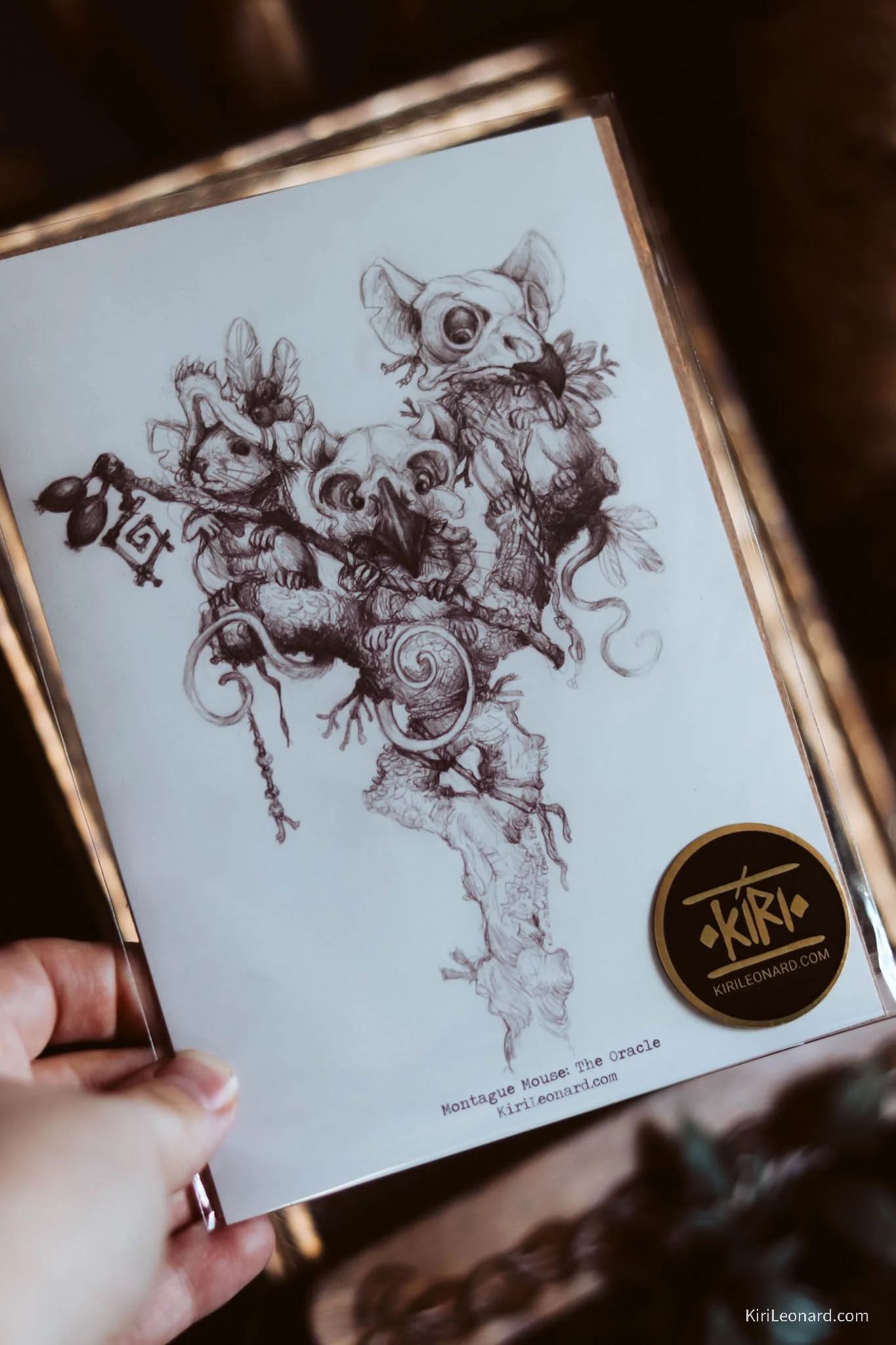 A hand holding an illustrated art print featuring a fantasy creature with a mouse head, bird skull, cat, and other animal parts, arranged in a tree-like formation with intricate details and shading.