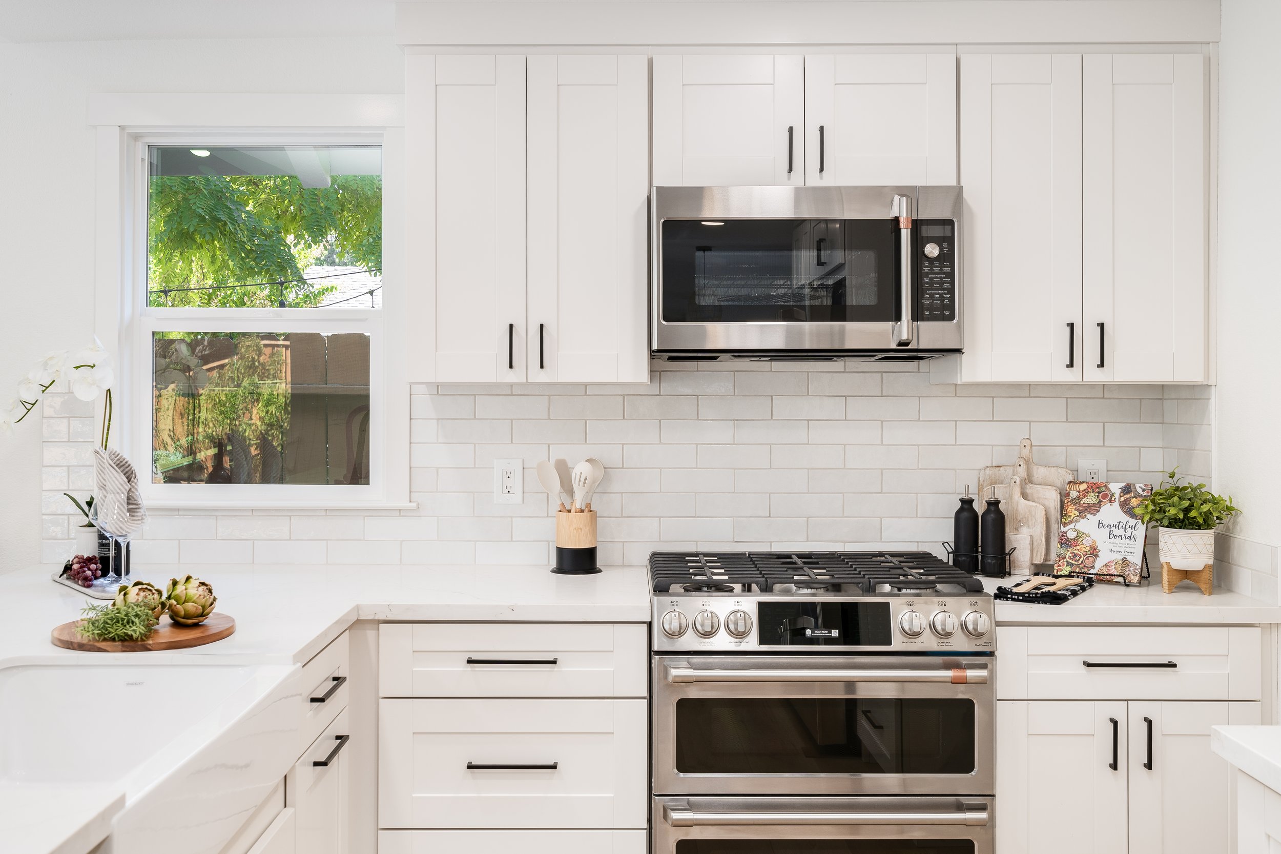 Modern kitchen with white cabinets, stainless steel oven and microwave, white subway tile backsplash, and a countertop with decor including a cutting board with artichokes, utensils in a holder, and potted plant.