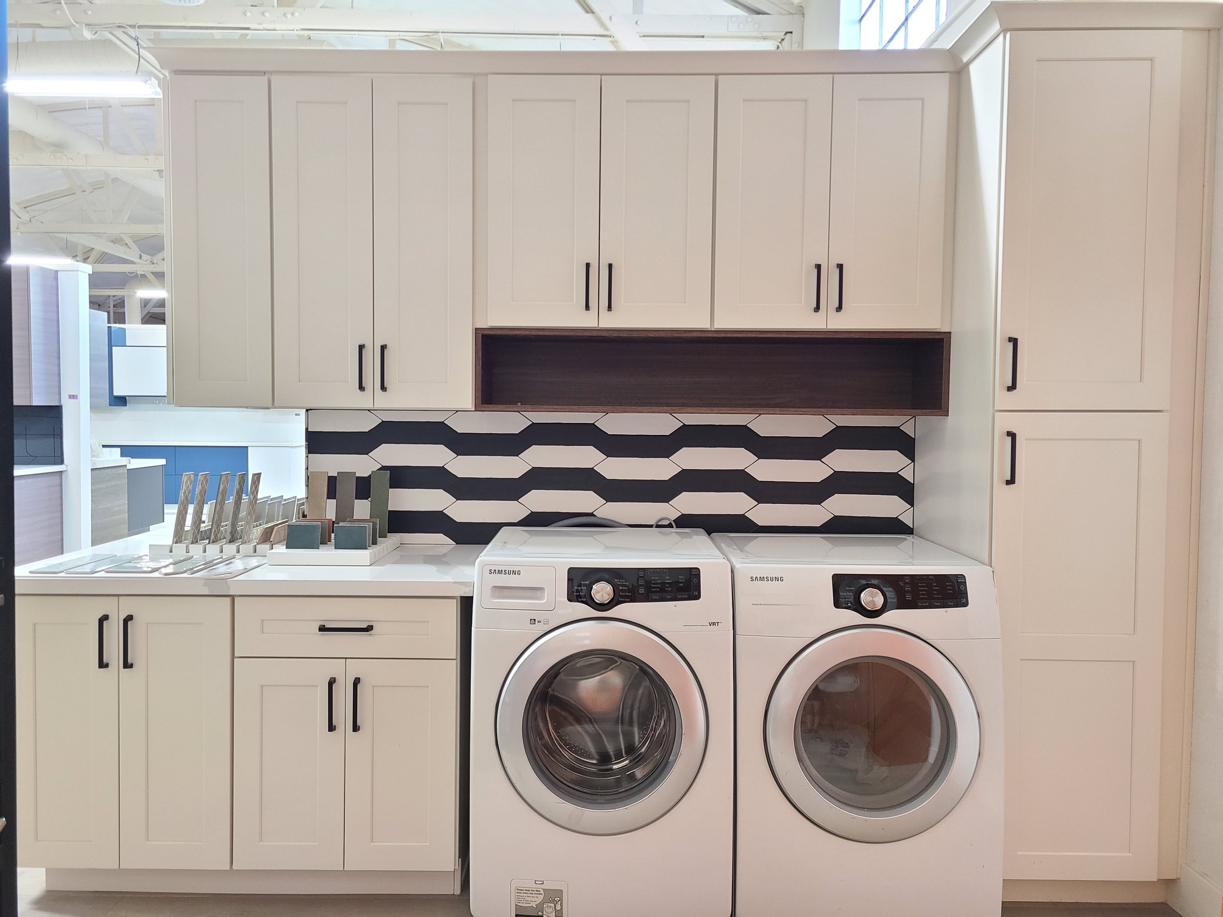 Laundry room with white cabinets, black handles, Samsung washer and dryer, and geometric tile backsplash.