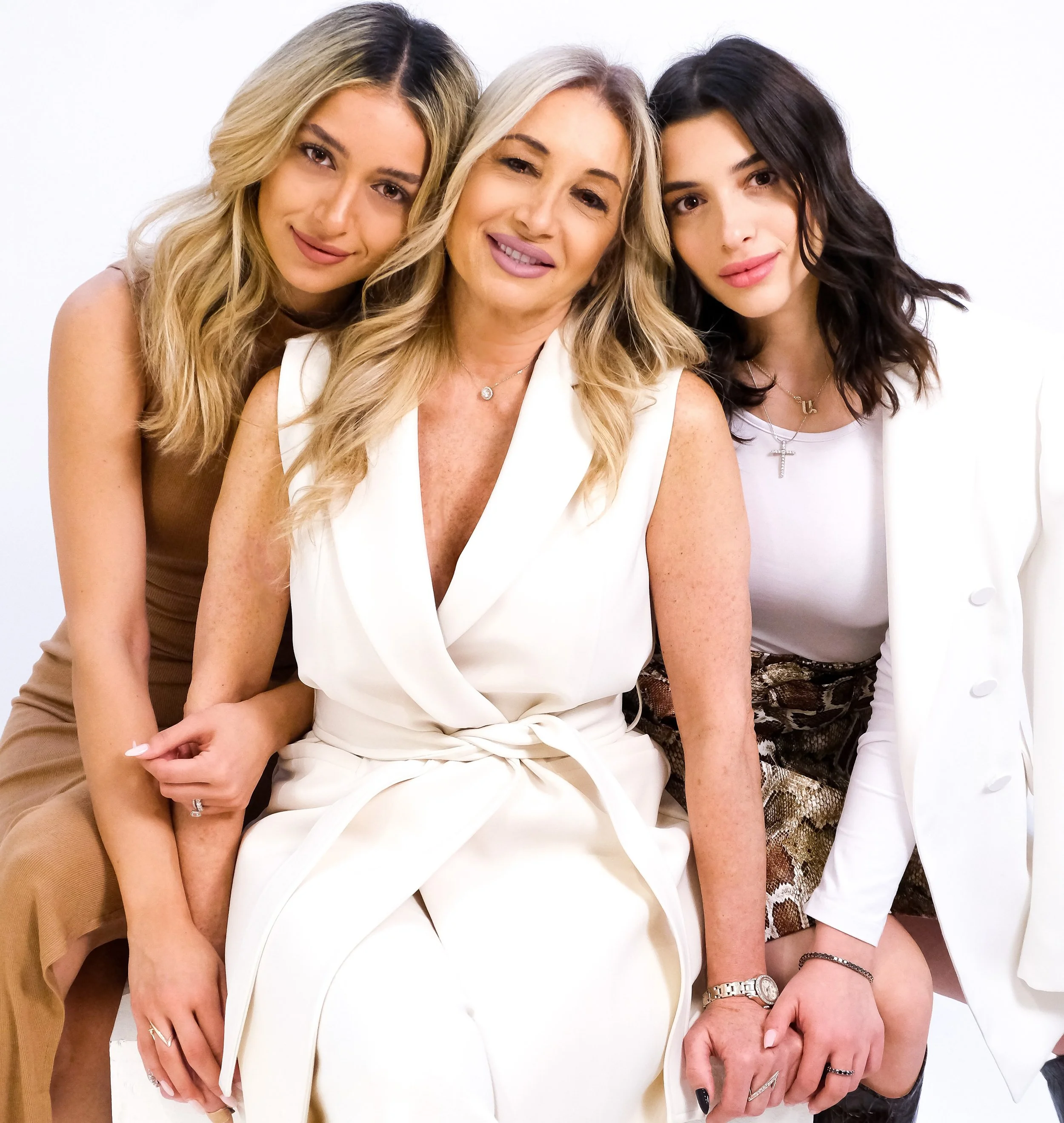 Three women sitting close together, smiling, with one woman in the center wearing a white sleeveless outfit, and two women on either side wearing casual stylish outfits.