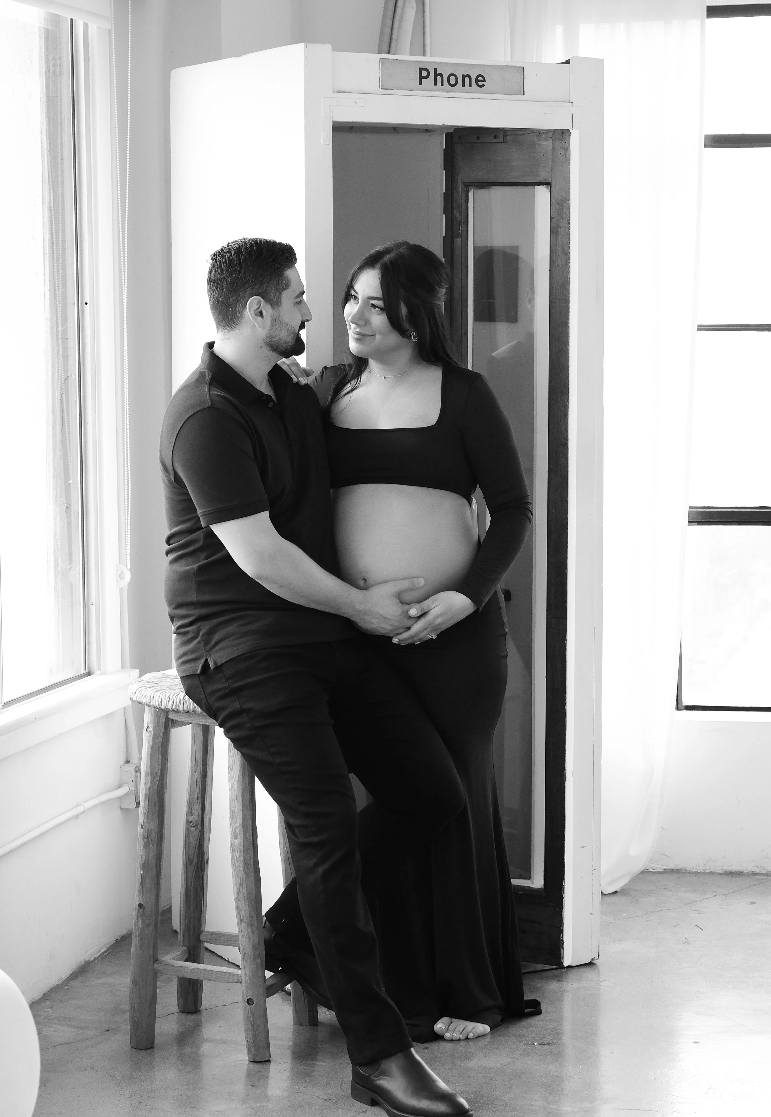Black and white photo of a pregnant woman standing inside a vintage phone booth, gently holding her belly, with a man sitting on a high stool next to her, both gazing at each other affectionately.