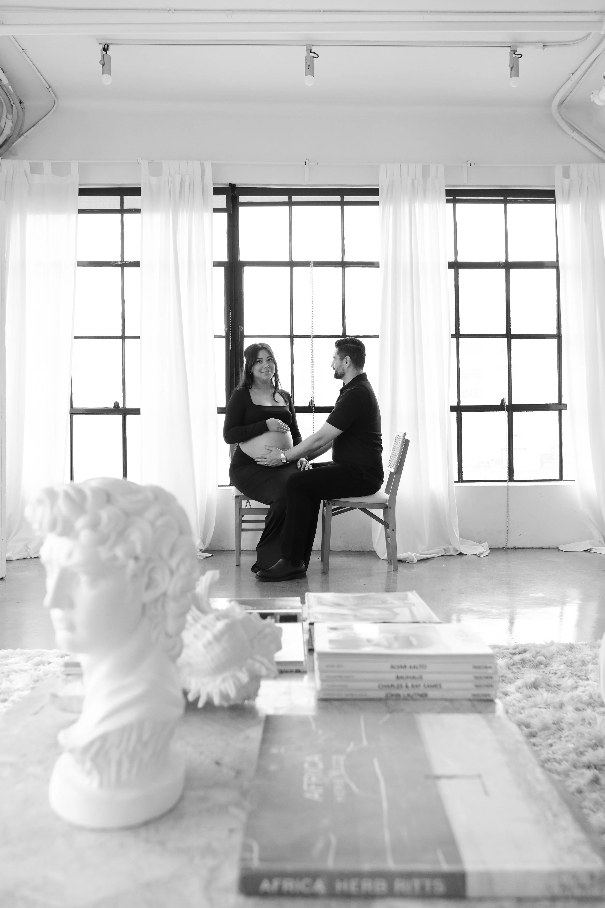 A couple in a studio with large windows and white curtains, the pregnant woman and man sitting on chairs, holding hands, with a white bust sculpture and a stack of books in the foreground.