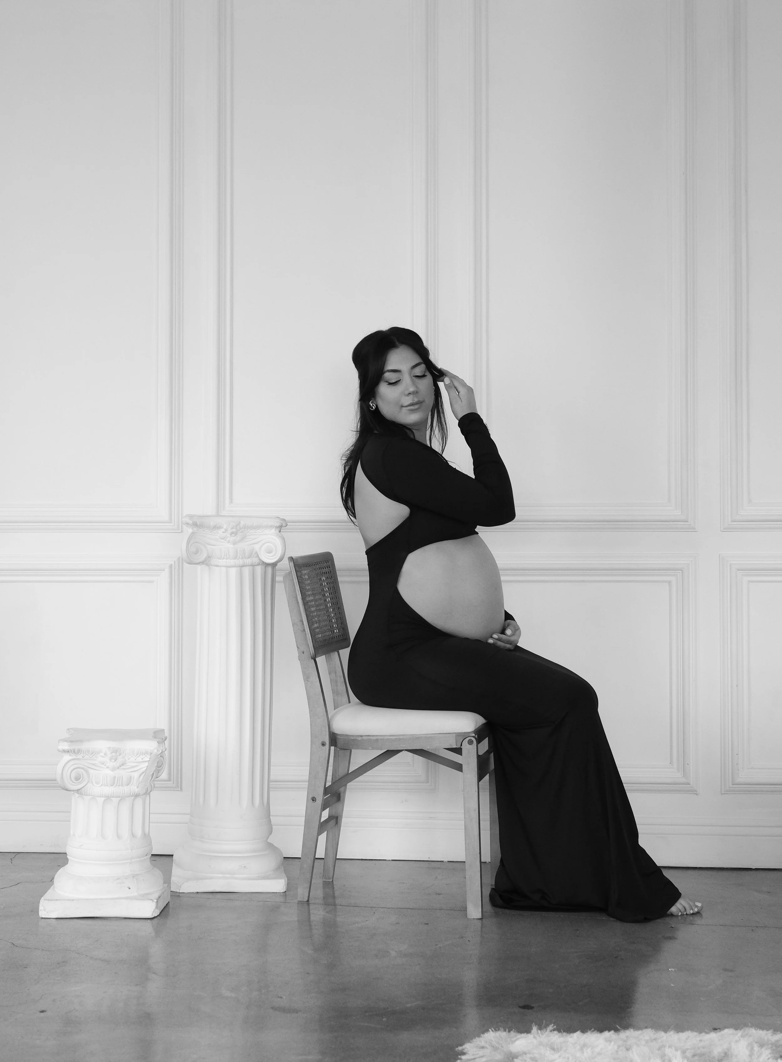 A pregnant woman with long dark hair, sitting on a chair in a black dress with cut-out sides, touching her belly, in a minimalist room with white paneling and a wooden floor.