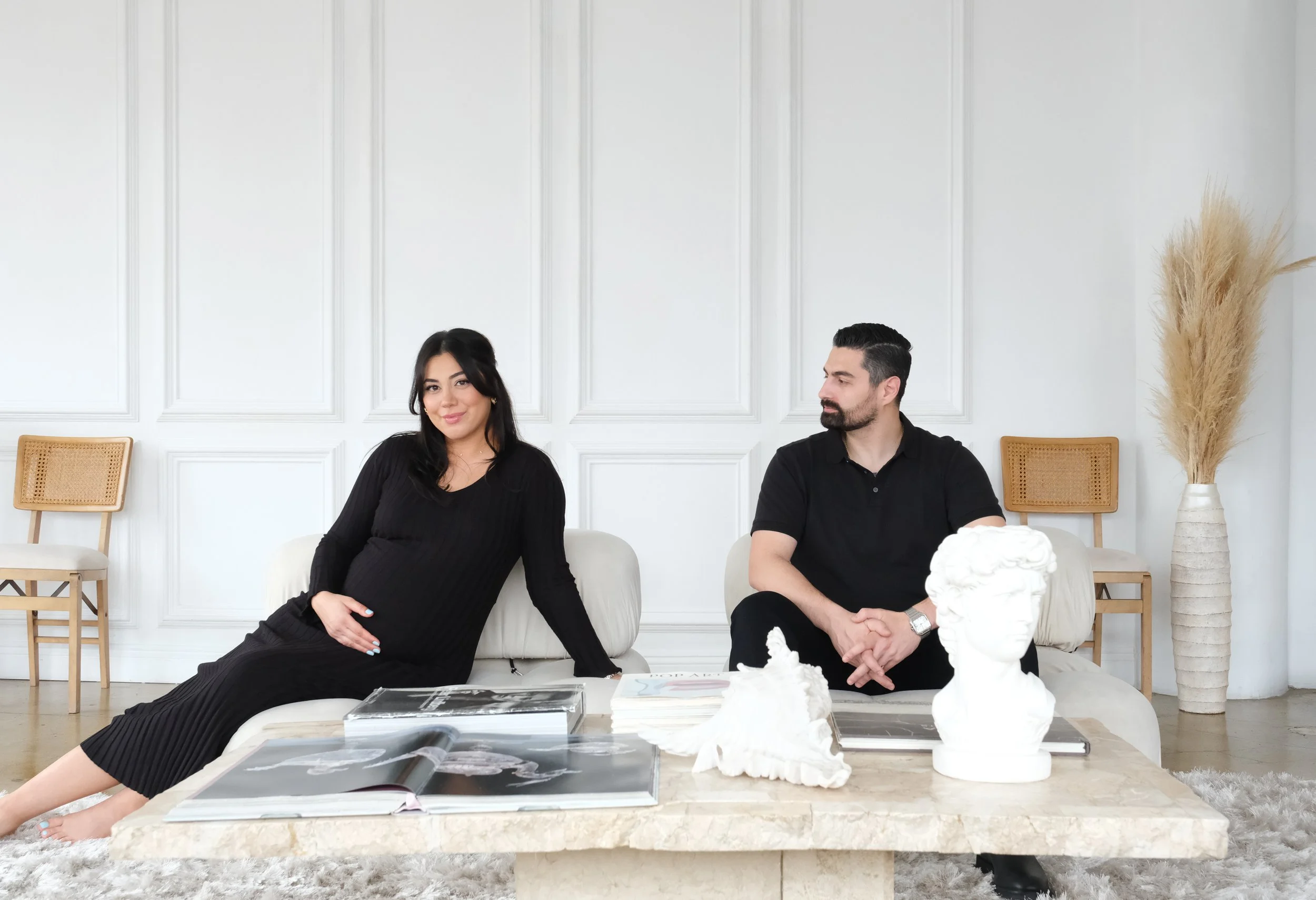 A woman in a black dress sitting on a white sofa cradles her pregnant belly while a man in a black polo shirt sits on the other side, both are in a modern, minimalist living room with white walls, wooden chairs, and a decorative vase with tall pampas