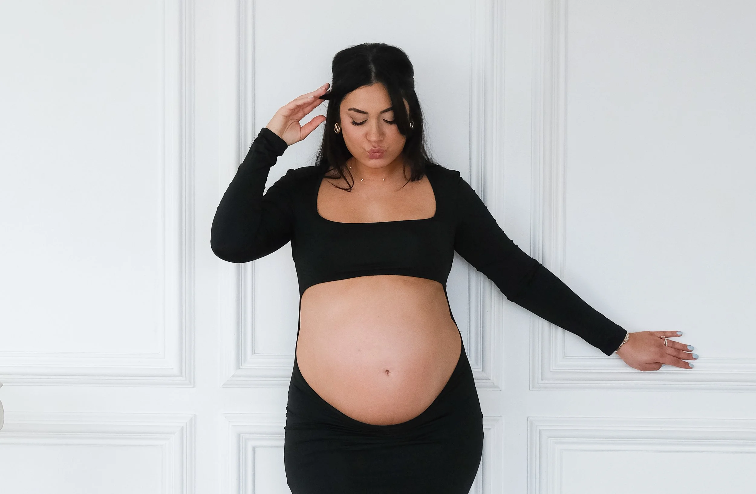 Pregnant woman wearing a black crop top and skirt, standing against white paneled wall, with one hand on her belly and the other touching her hair.