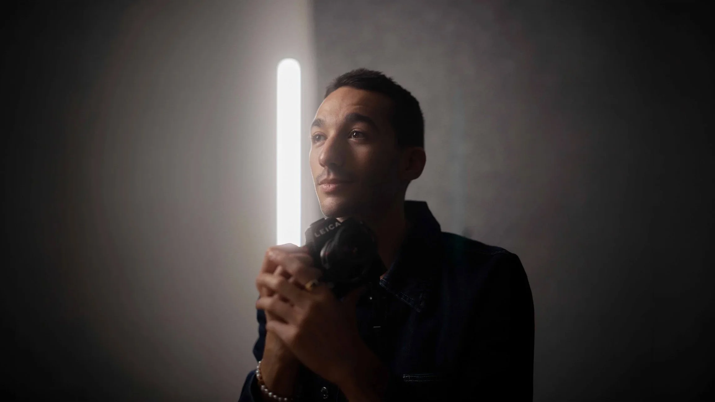 A man holding a camera with a bright vertical light in the background, partially facing the camera, with a dark background.