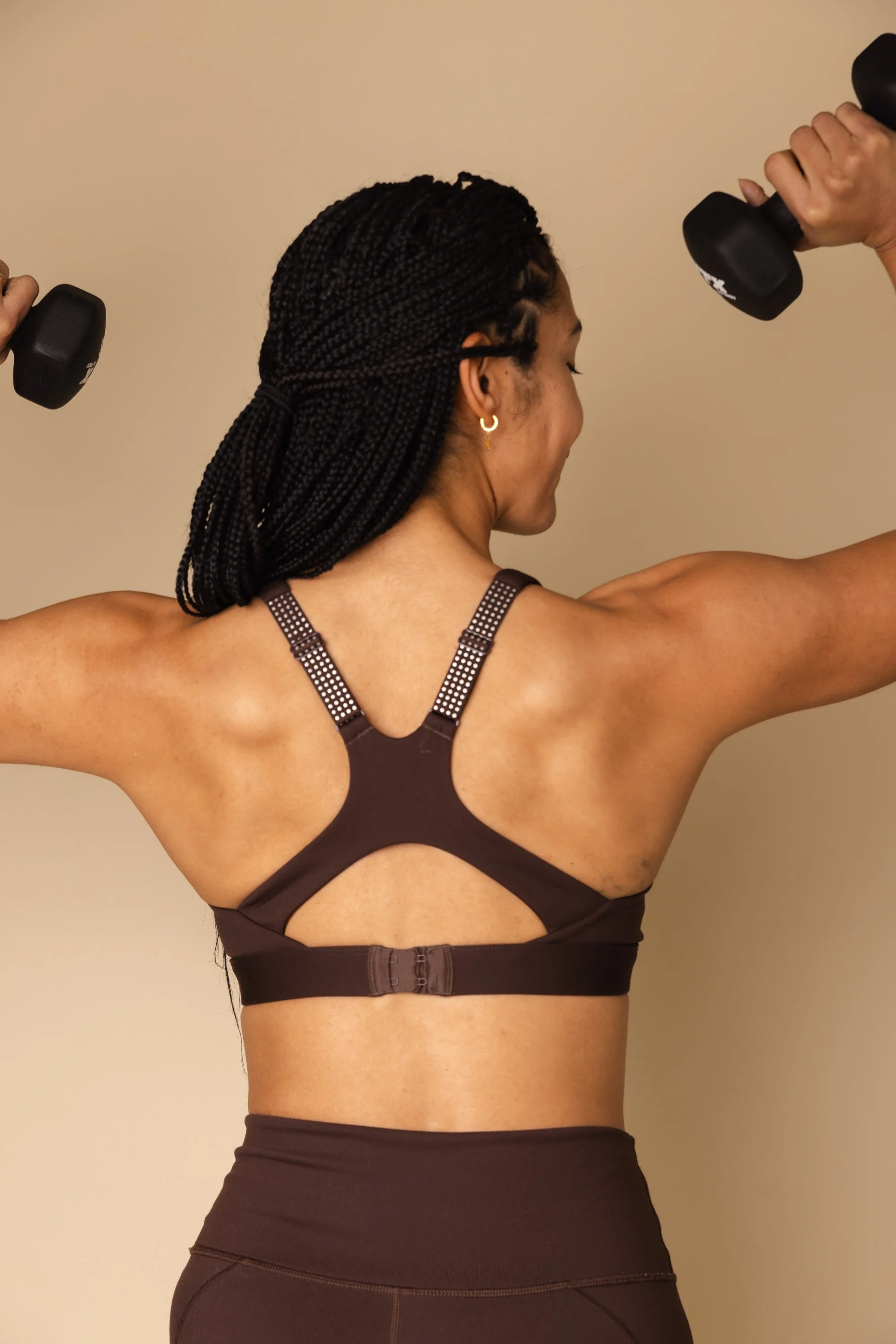 A woman with black braided hair, wearing a dark brown sports bra with a unique back design, holding black dumbbells in each hand, raised at shoulder height, performing shoulder exercises against a plain beige wall.