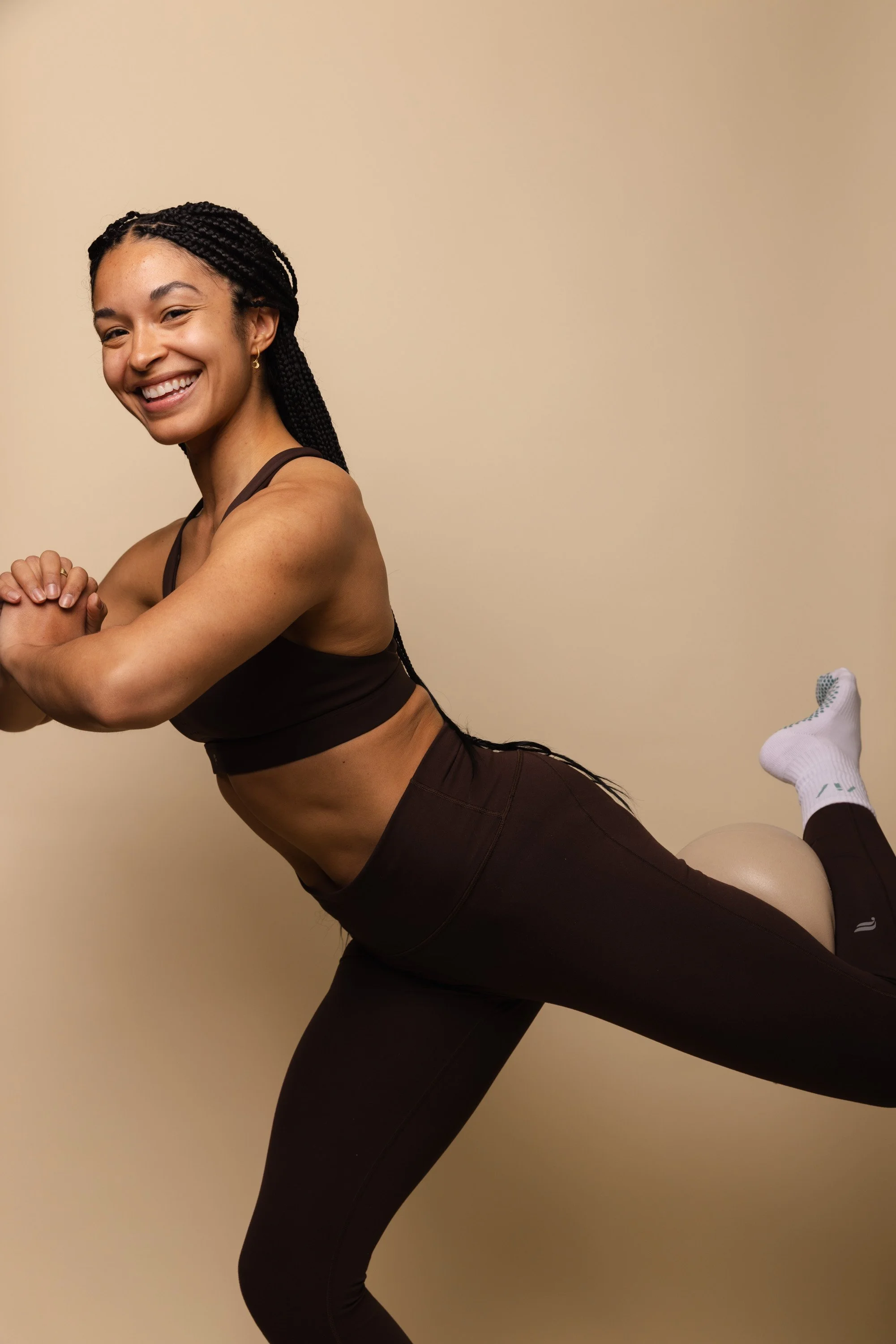 A woman smiling and doing a yoga pose against a plain beige wall. She is wearing a brown sports bra and matching leggings, and has her hair styled in long braids.
