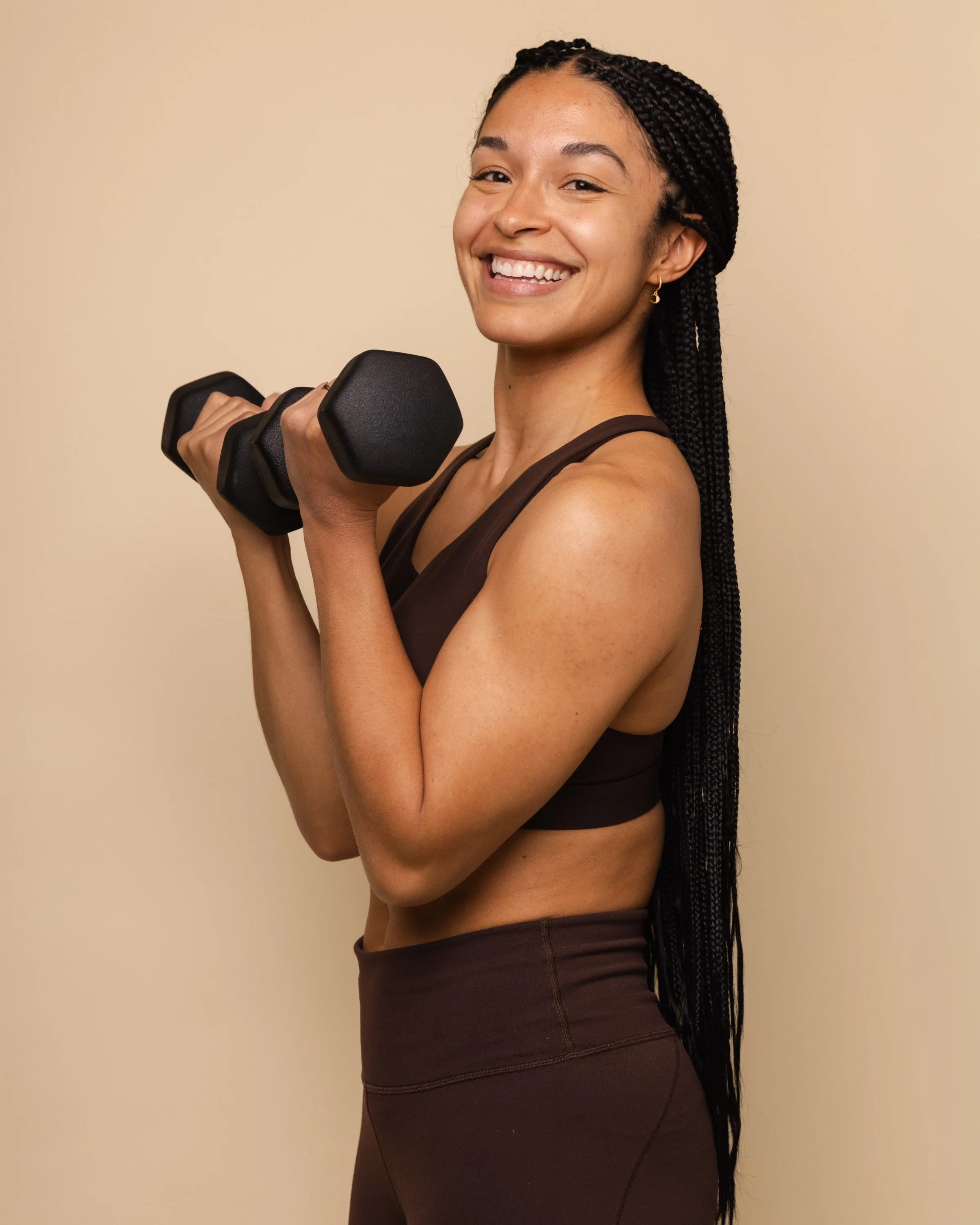 A woman smiling and holding a black dumbbell in her right hand, wearing a brown sports bra and matching workout leggings, with long braided hair, standing against a beige background.