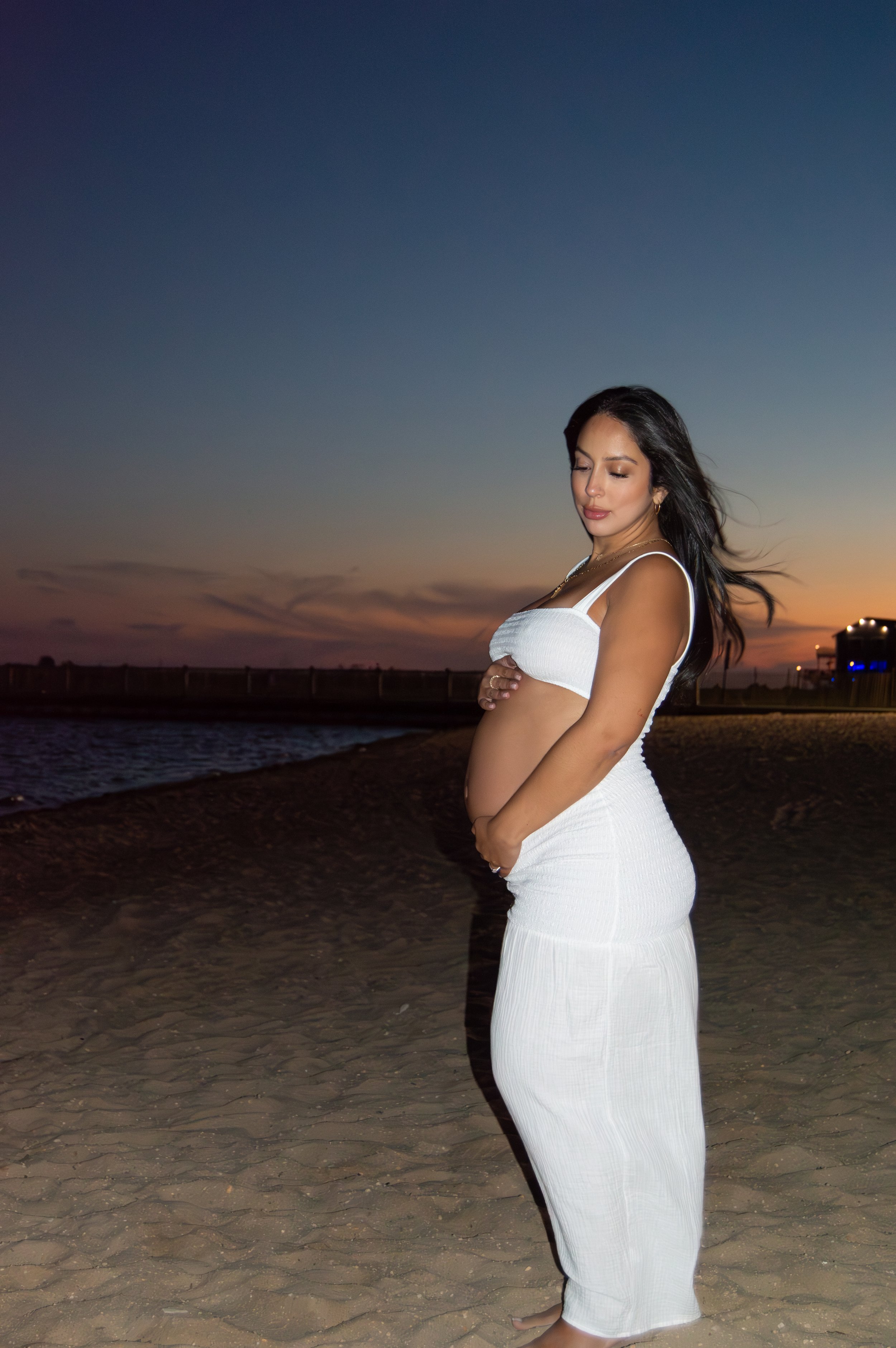 A pregnant woman in a white dress standing on a sandy beach at sunset, looking down at her belly with her hands clasped over her stomach.