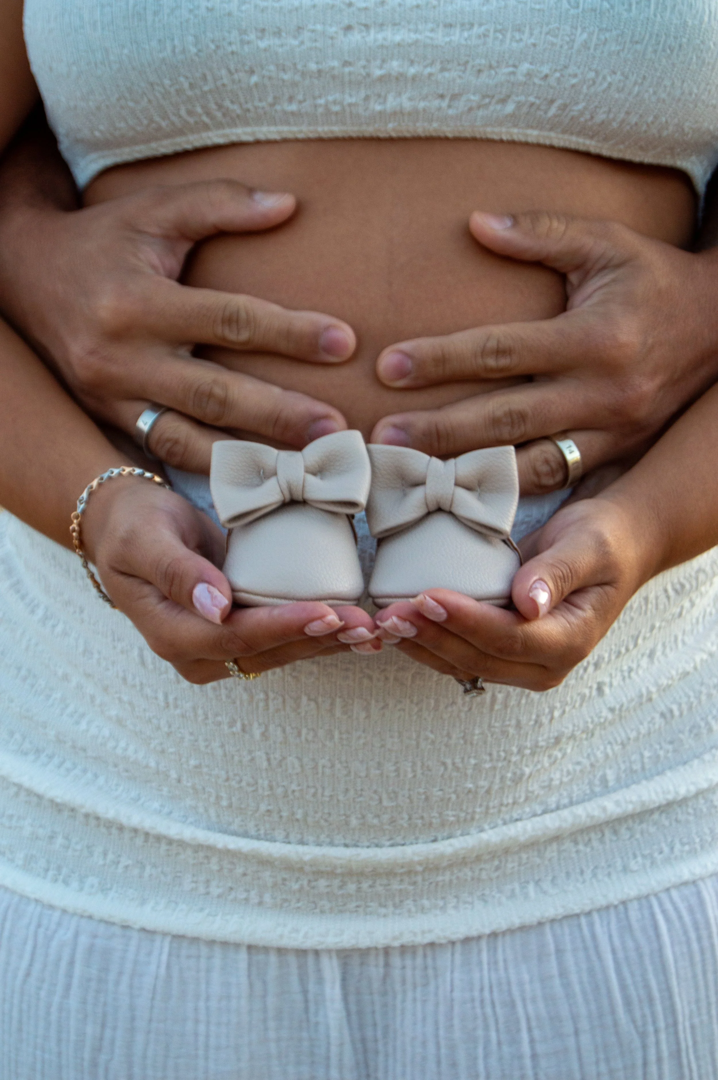 Close-up of a pregnant woman's belly with hands gently holding it, and a pair of small beige baby shoes with bows placed on her stomach.