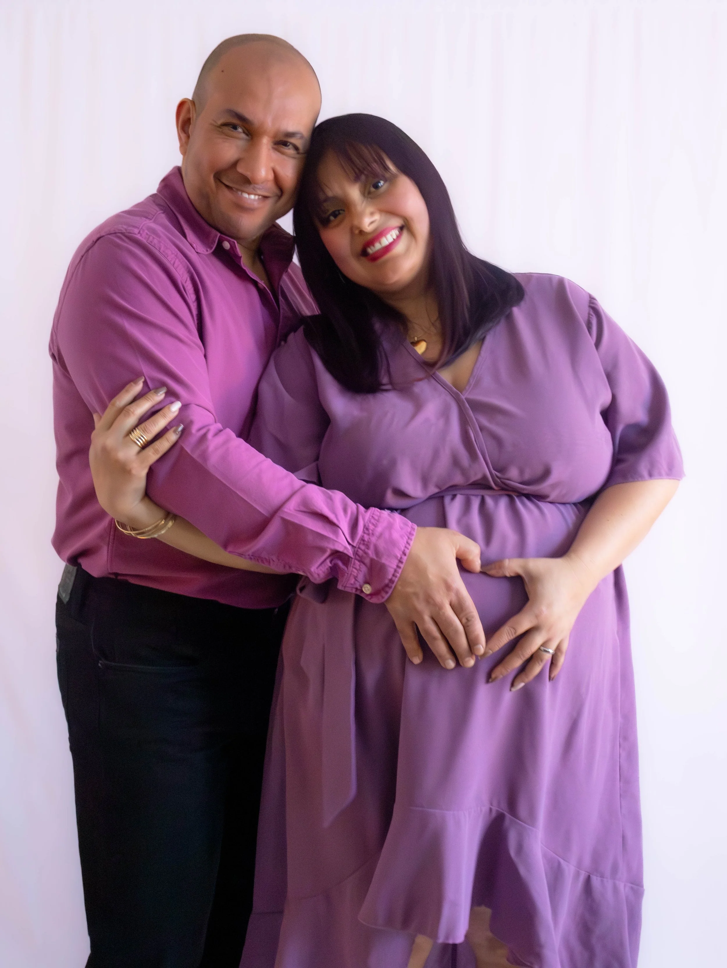 A happy couple, dressed in purple, embraces each other and touches the pregnant woman's belly, standing against a white background.