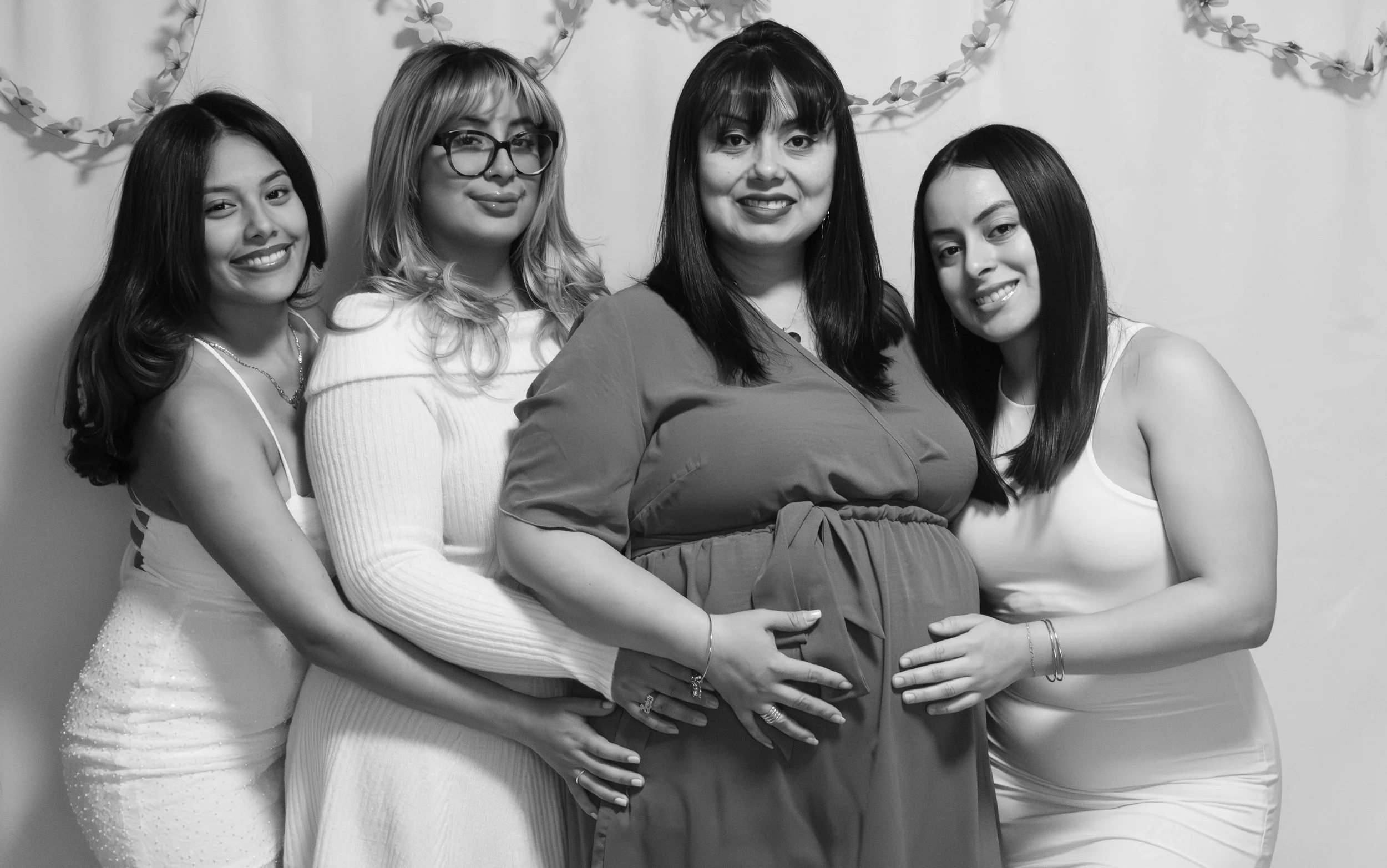 Four women standing close together, with one visibly pregnant, smiling at the camera in front of a decorated backdrop.