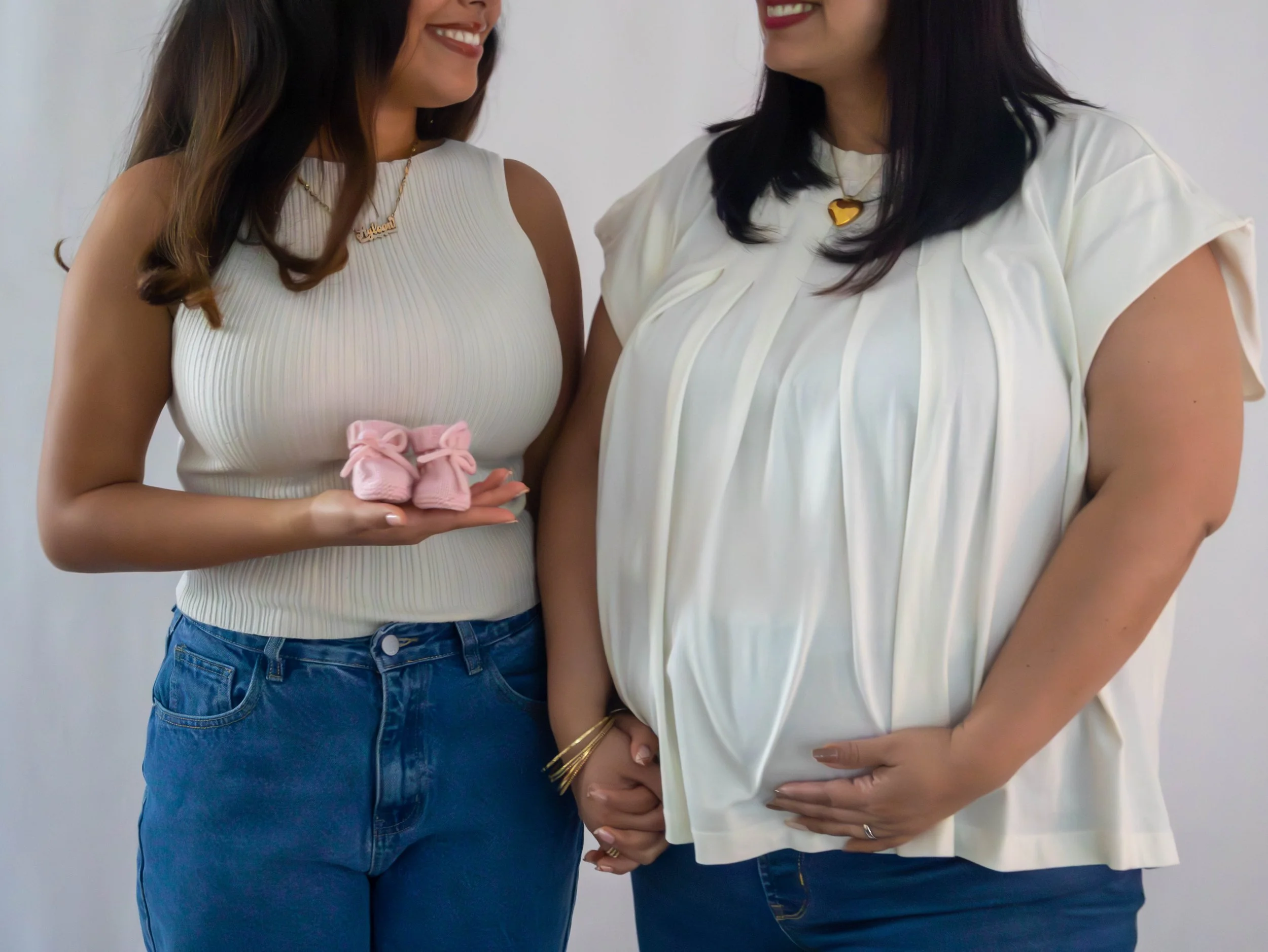 Two women standing together, one holding pink baby shoes, the other with hands clasped, both smiling and wearing light-colored tops.