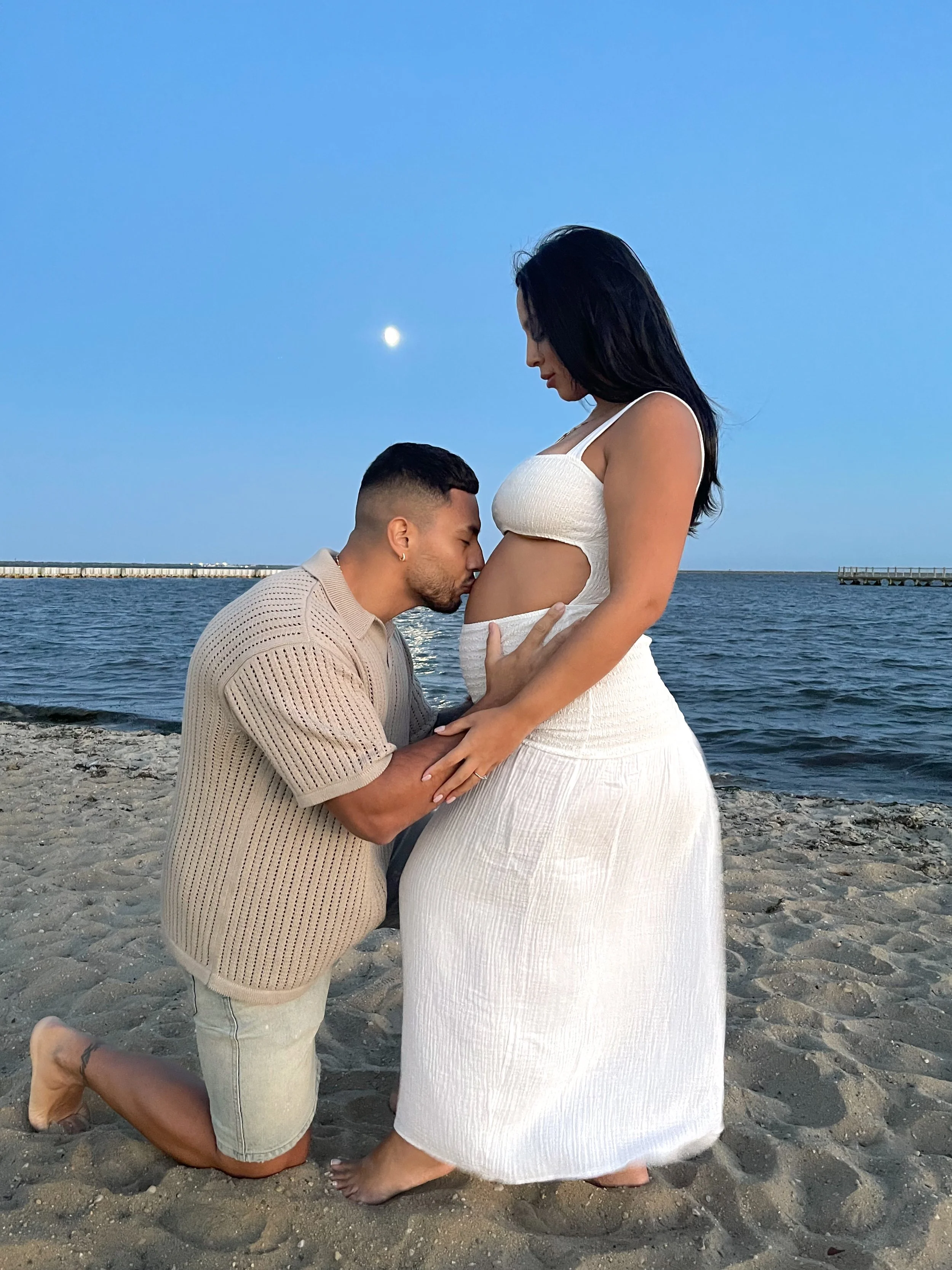 A couple on the beach, with the man kneeling and kissing the pregnant woman's belly, and the woman standing in a white dress holding her belly, during twilight with the moon in the sky.