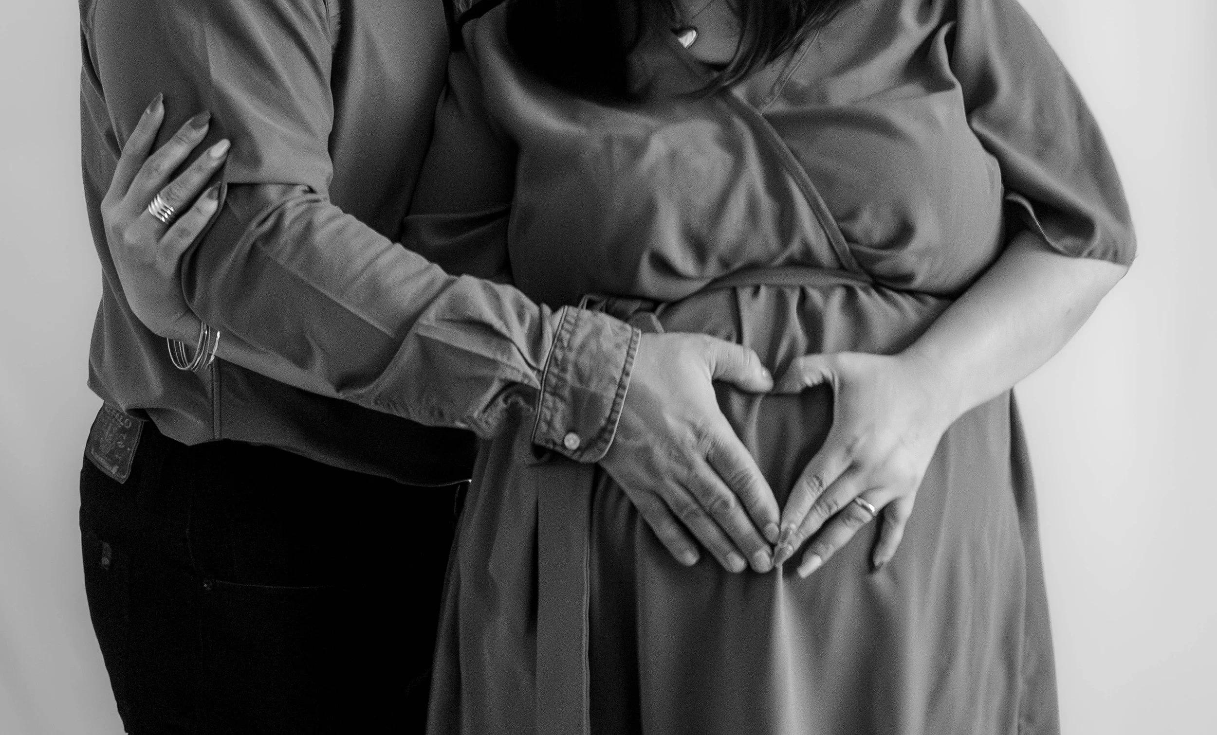 Two women forming a heart shape with their hands on her pregnant belly, one woman hugging the other around the shoulders.