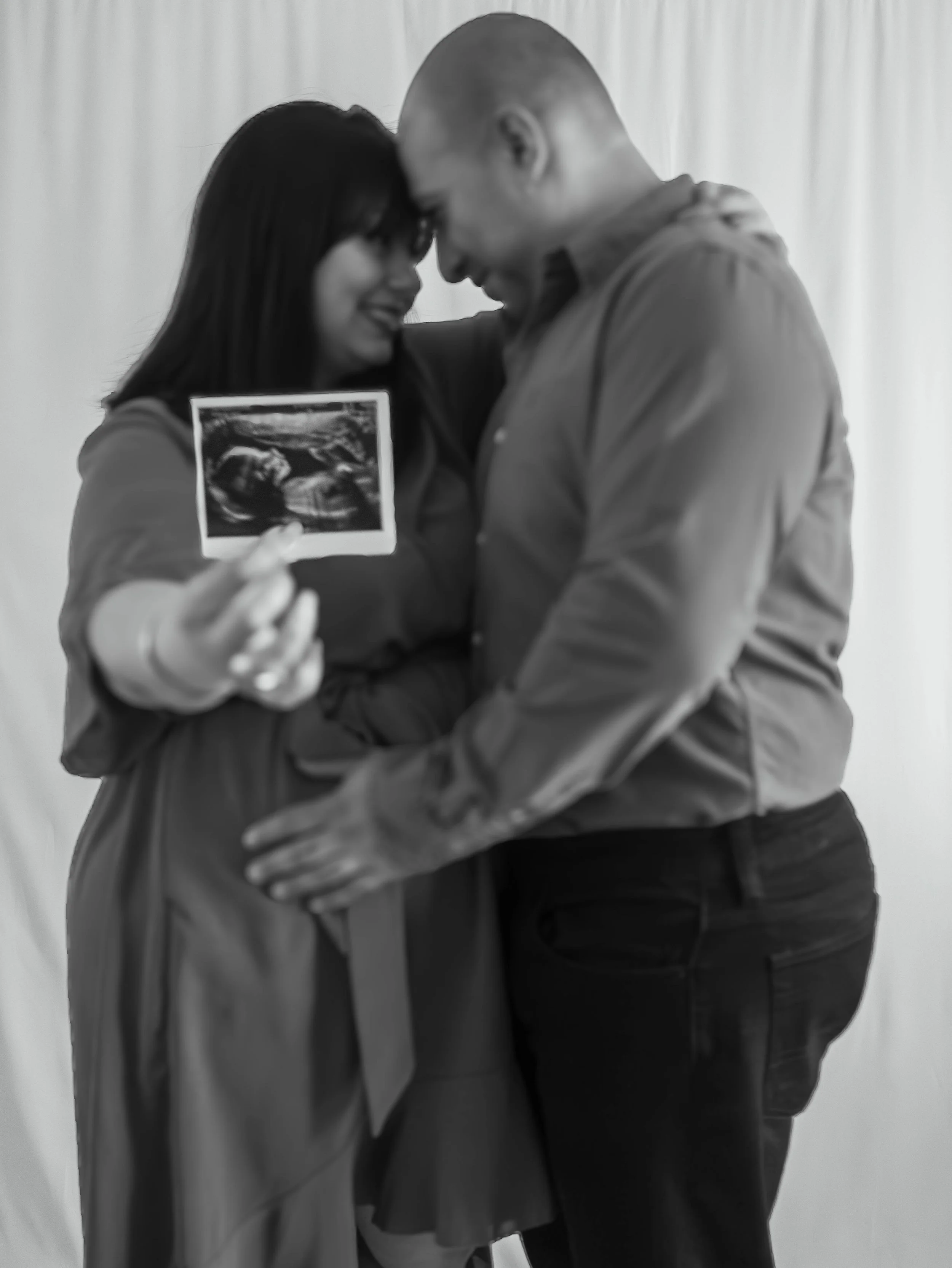 A couple holding an ultrasound picture, smiling and touching foreheads in front of a plain background.