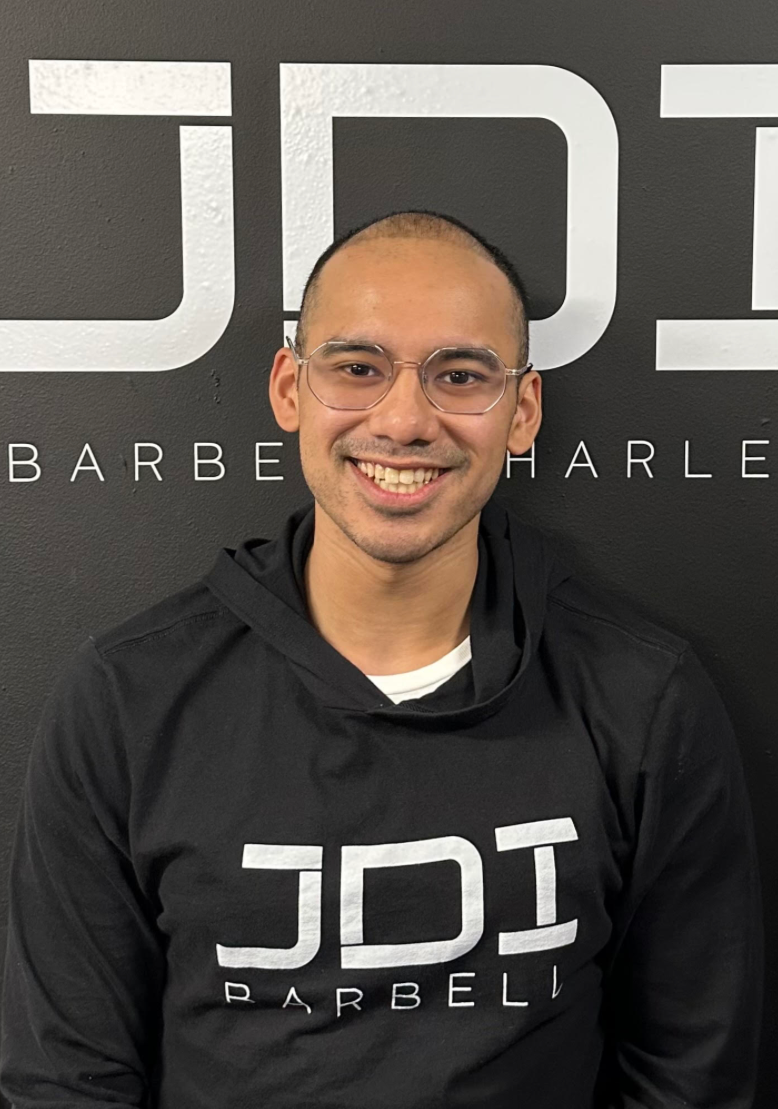 A smiling man with glasses, short hair, and a black hoodie sitting in front of a black wall with large white text that reads "JDI" and smaller text that reads "Barber Harlem."