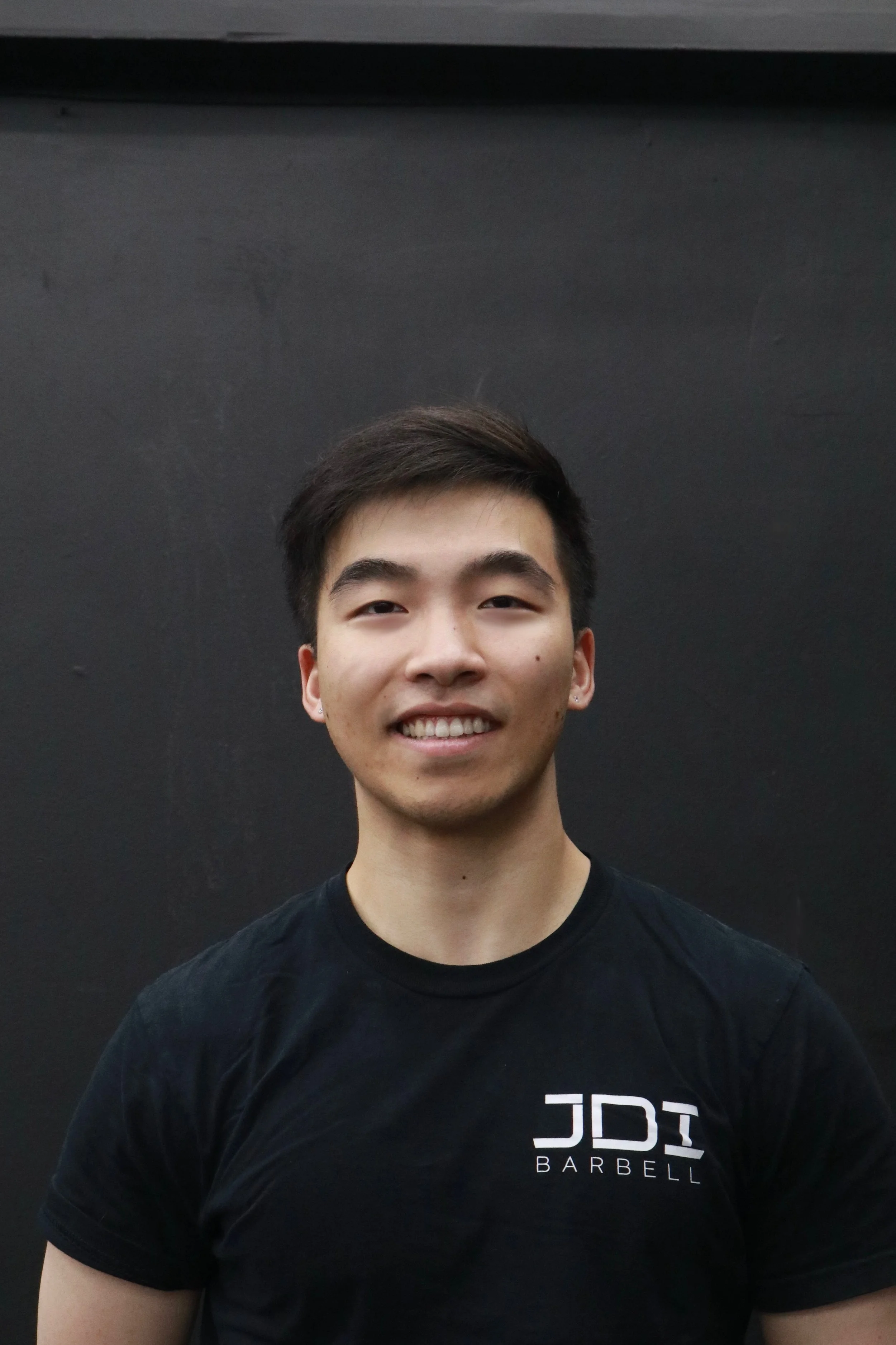 A young man with short dark hair smiling in front of a black background, wearing a black T-shirt with white text reading 'JDI BARBELL'.