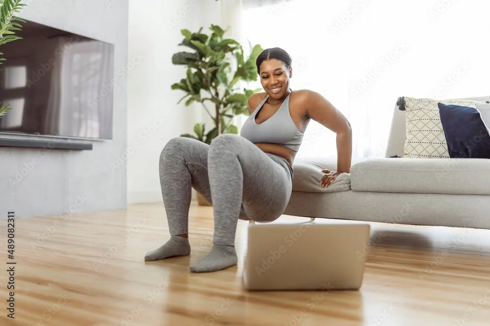 A Black woman in workout clothes doing sit-ups on the floor in a living room while looking at a laptop.