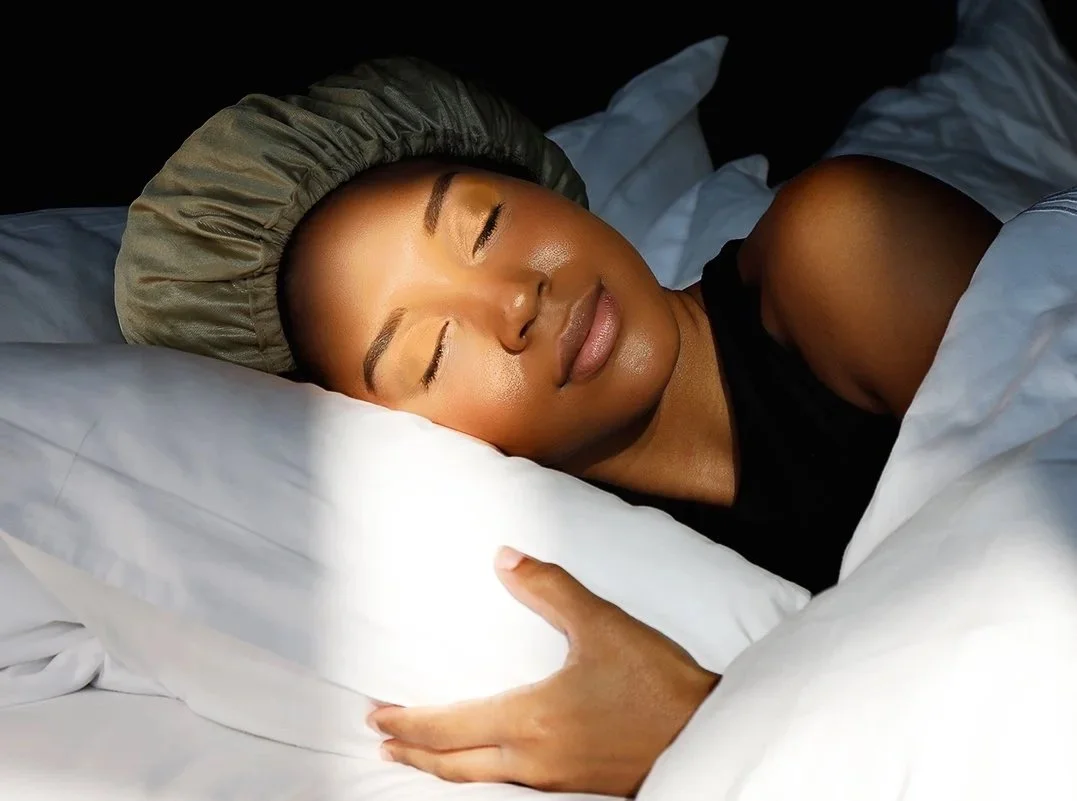 A Black woman with smooth skin and defined eyebrows sleeps peacefully on a pillow, wearing a sleep cap, under a blanket.