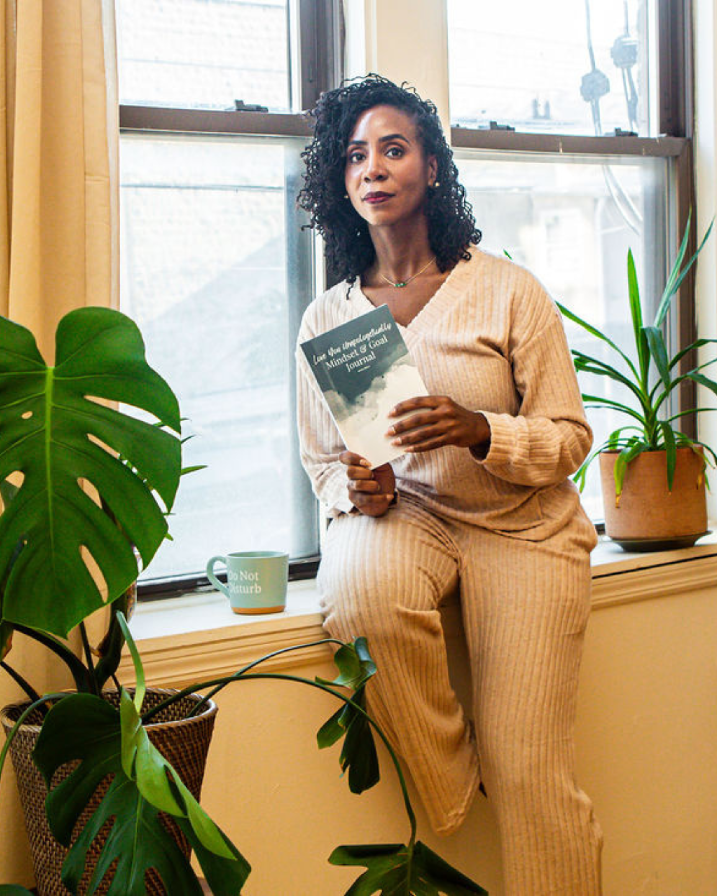 Woman with curly hair wearing beige ribbed sweater holding a magazine, standing near a window with yellow curtains and large green plants, with a mug on the windowsill.