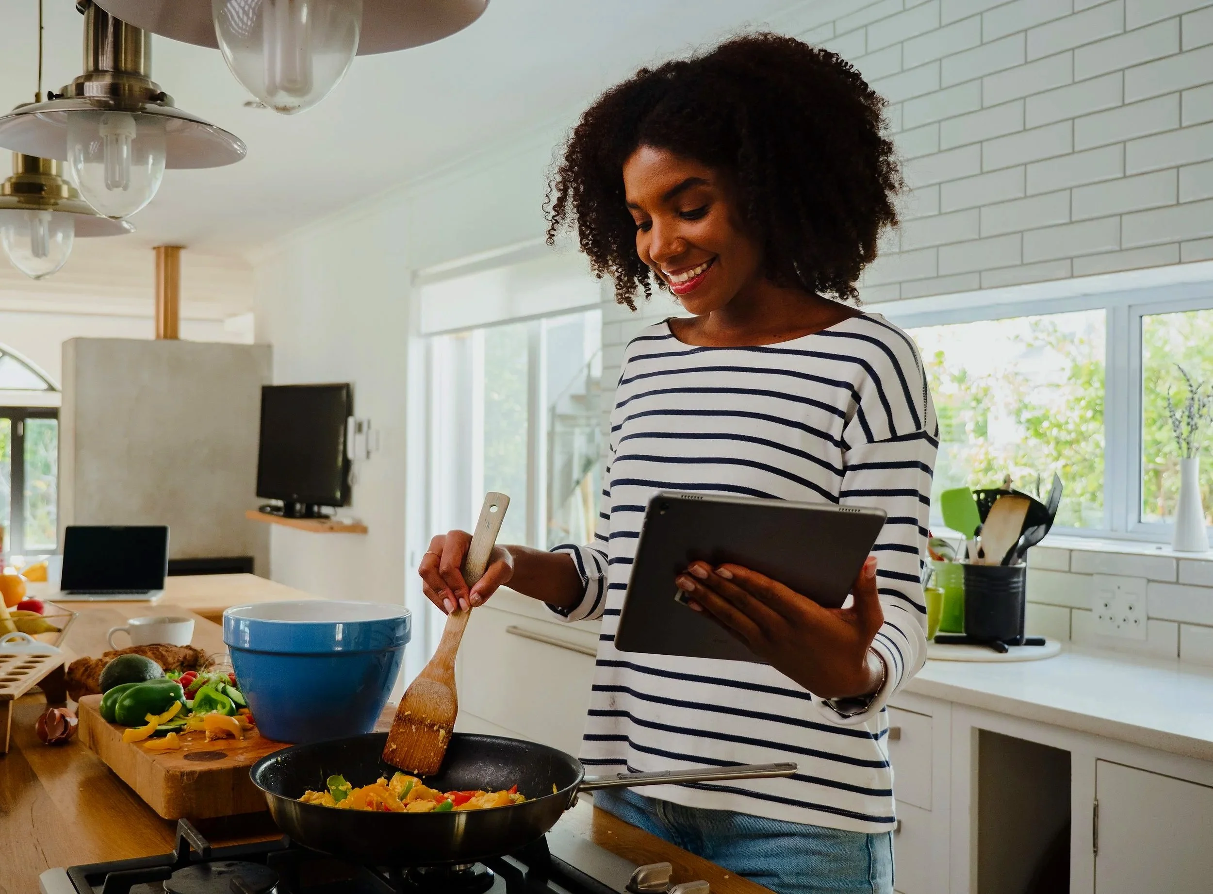 A woman with curly hair wearing a striped shirt is cooking in a modern kitchen. She is holding a wooden spoon and a tablet device, standing near a stove with a black skillet. The kitchen has a white brick wall, large windows, and various kitchen utensils and appliances on the counter.
