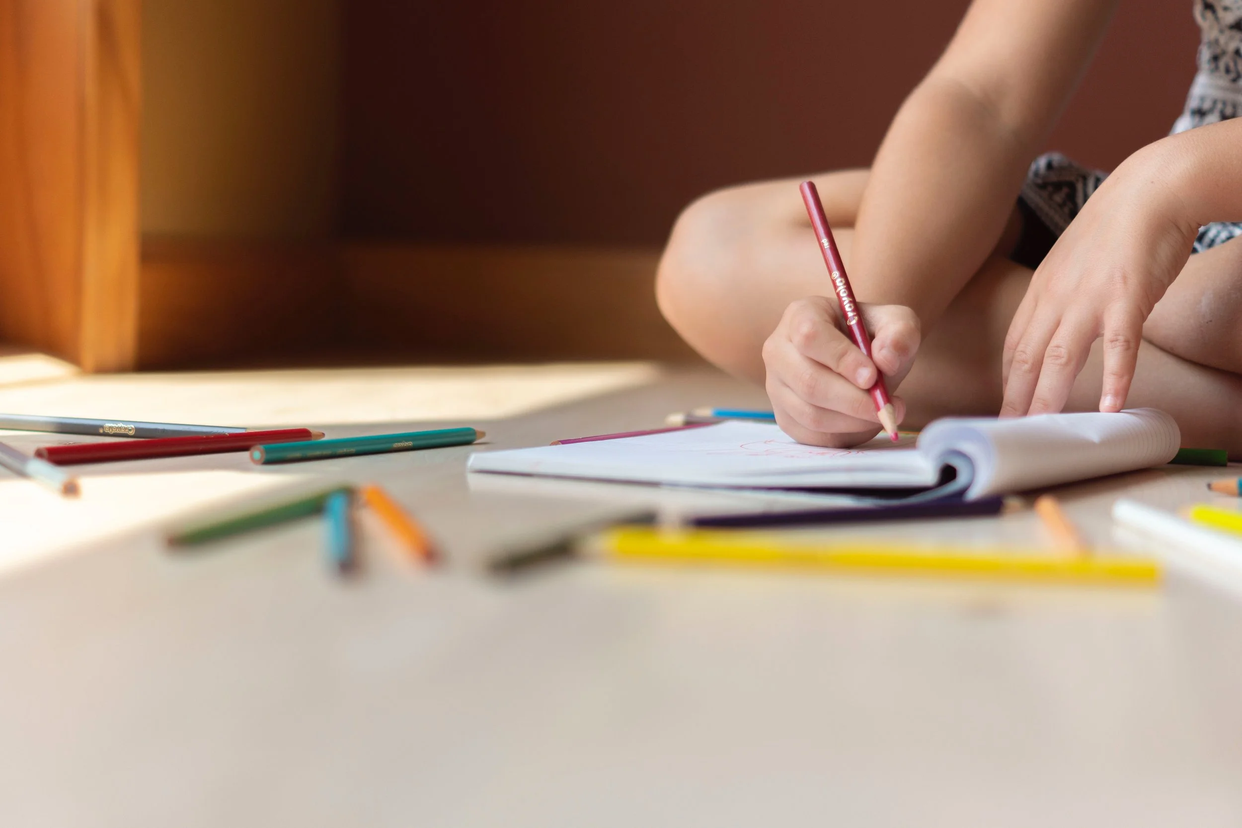 Child sitting on floor, drawing in a notebook with colored pencils scattered around.
