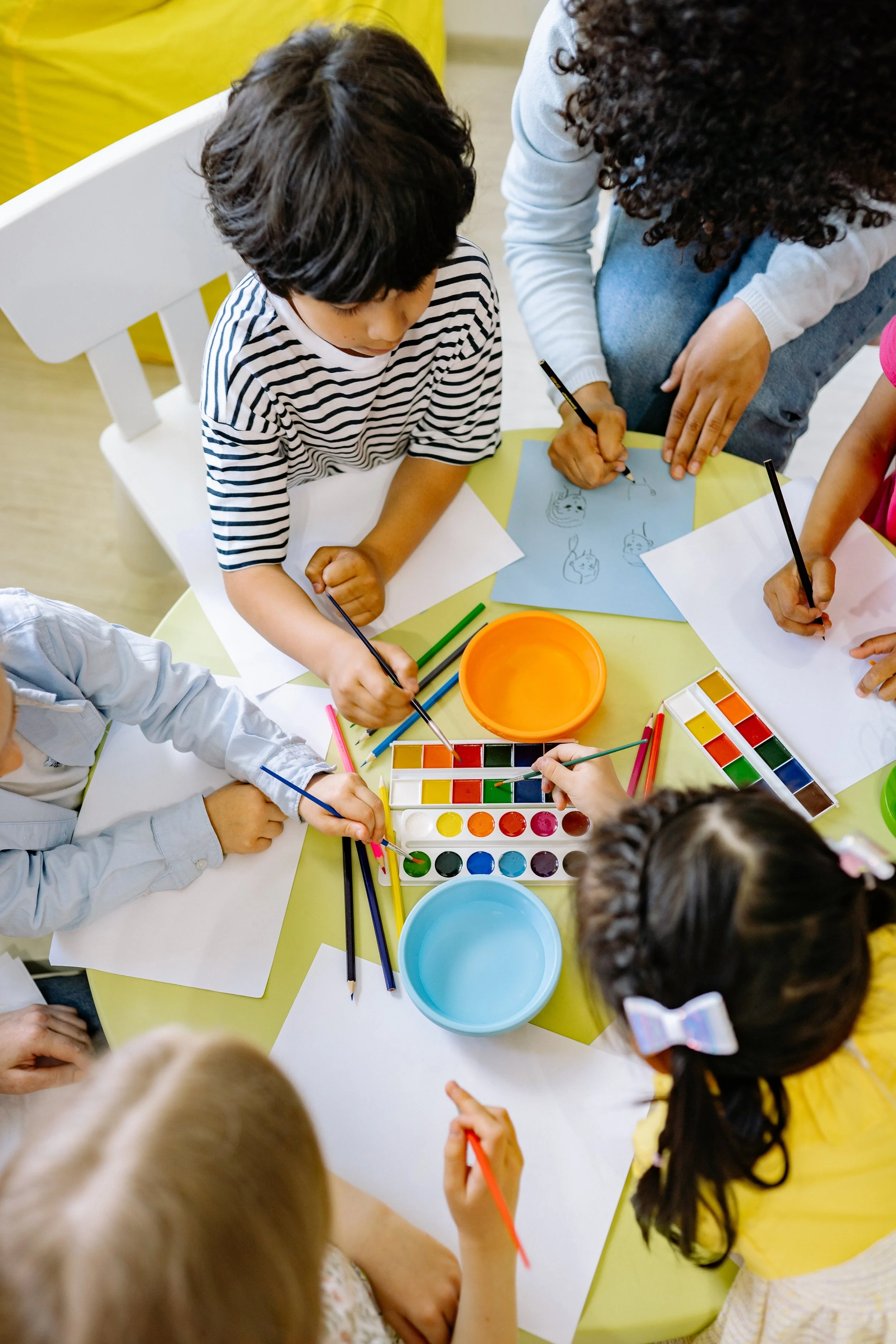 Children and an adult are gathered around a table painting and drawing with watercolors, surrounded by bowls of paint and sheets of paper.