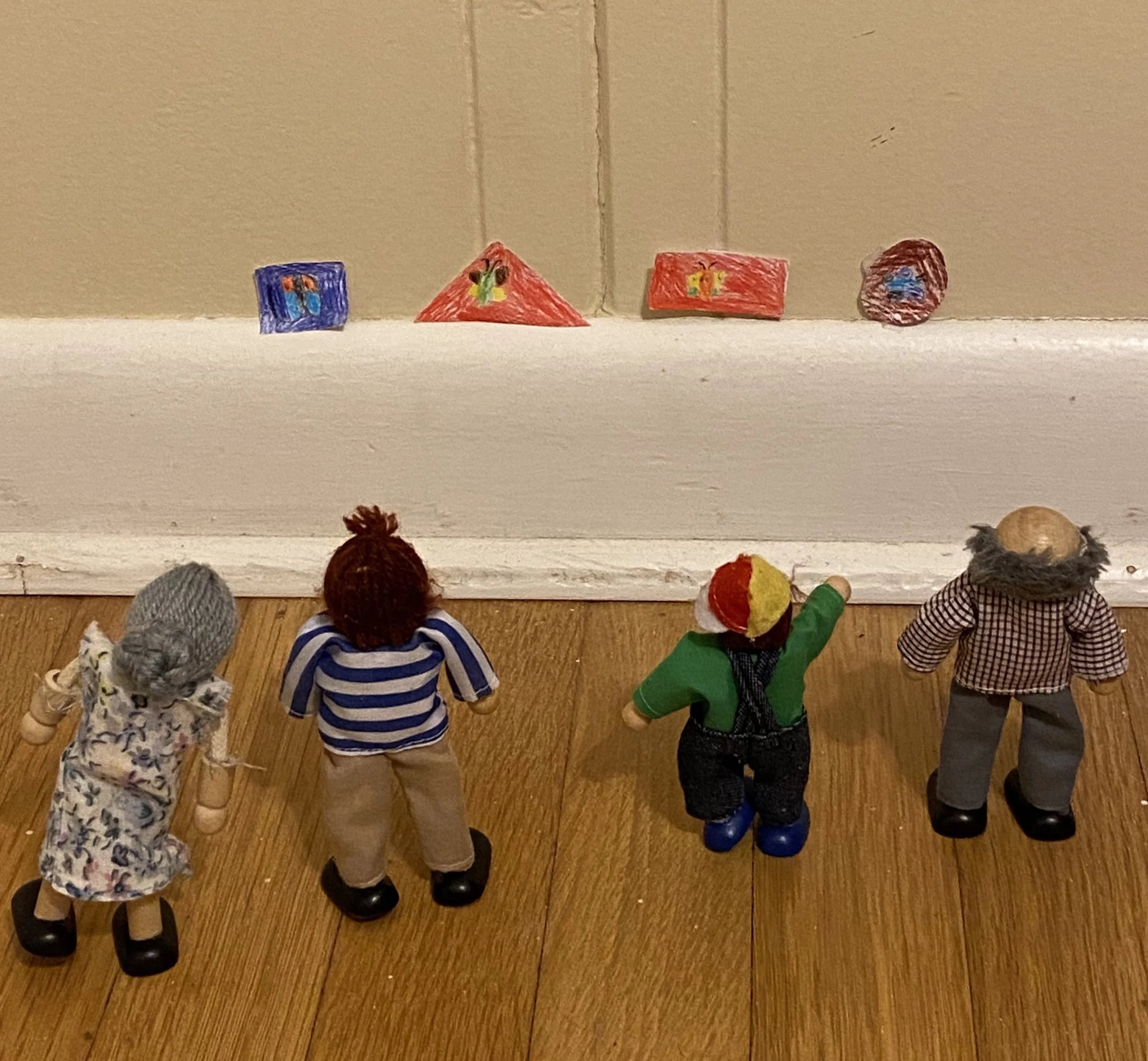Four doll figurines with various outfits and hairstyles stand on a wooden floor, facing a small collection of hand-drawn, colored paper boxes set on a white surface against a beige wall, resembling a miniature art display.