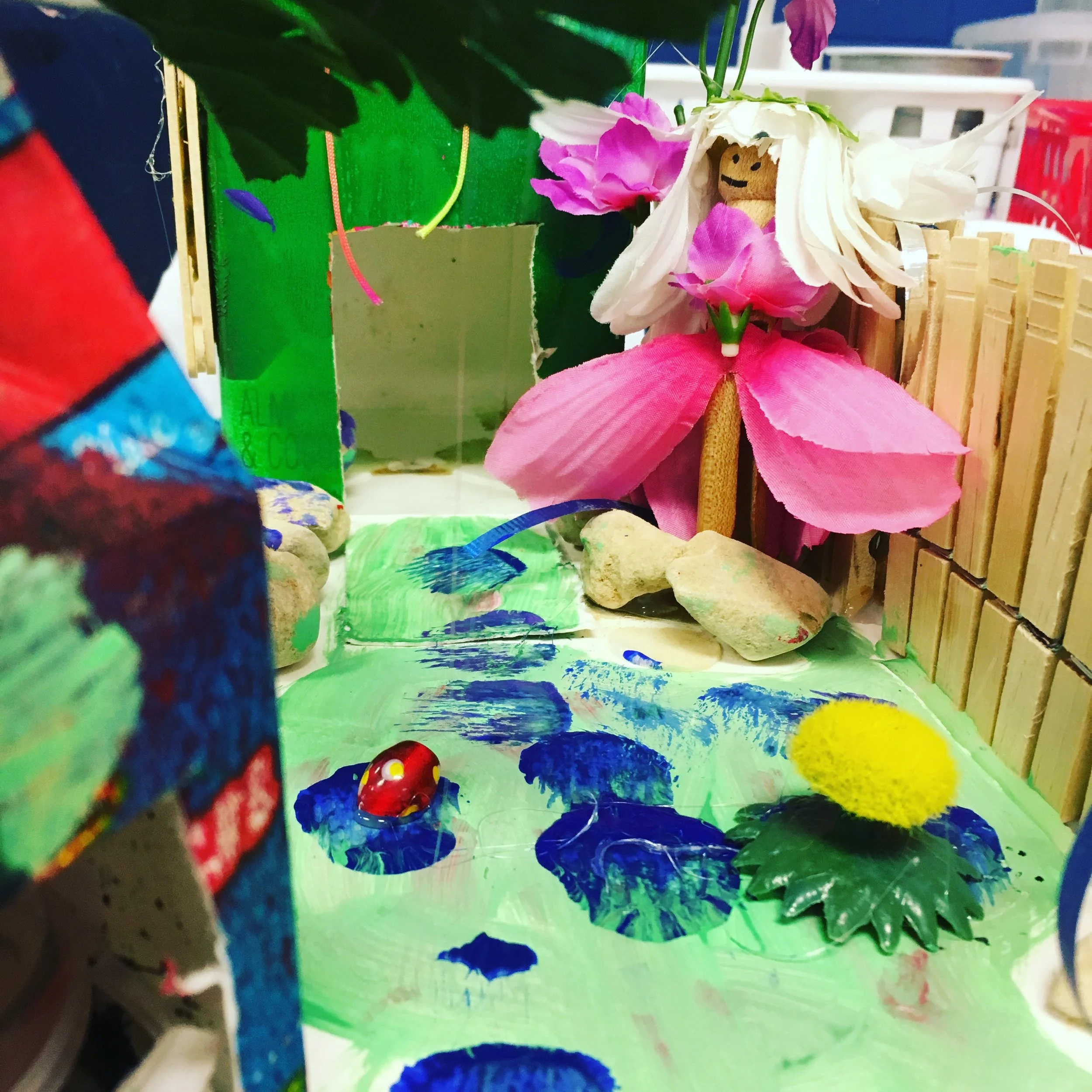 A miniature diorama scene of a fairy garden with a fairy figurine made of pink and white flowers, painted rocks, a small artificial pond with a ladybug figurine, and painted green and blue rocks, surrounded by a painted backdrop and various craft mat