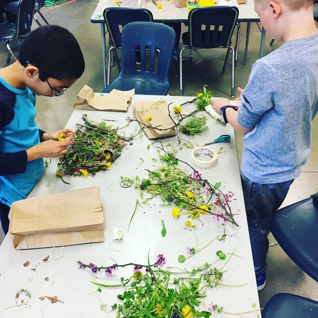 Two children working on a craft project with flowers, leaves, and twigs on a table. One child is on the left, wearing glasses and a blue shirt, arranging flowers. The other child is on the right, wearing a gray shirt, holding a bunch of flowers. The 