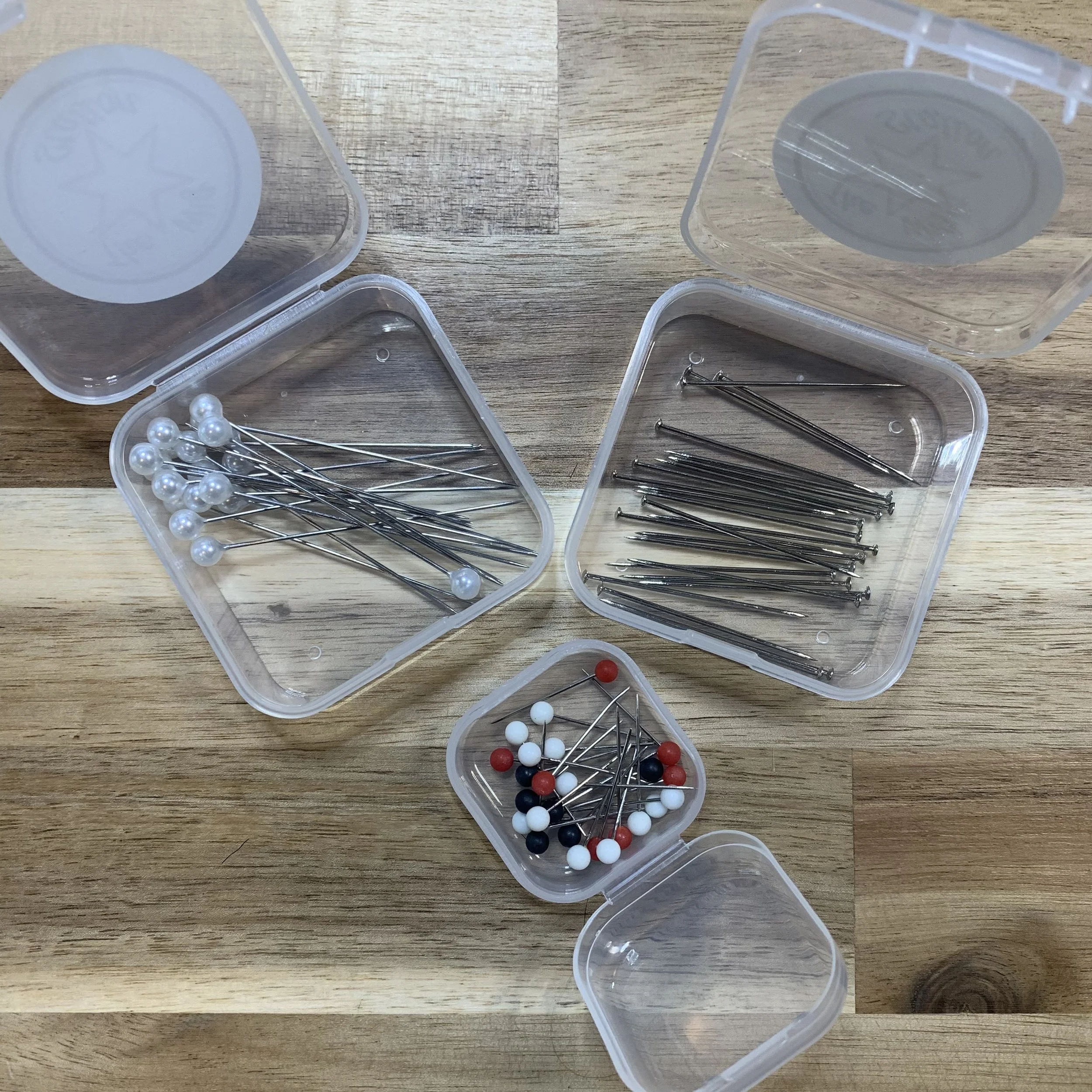 Multiple plastic containers with sewing pins, some with white or multicolored heads, on a wooden surface.