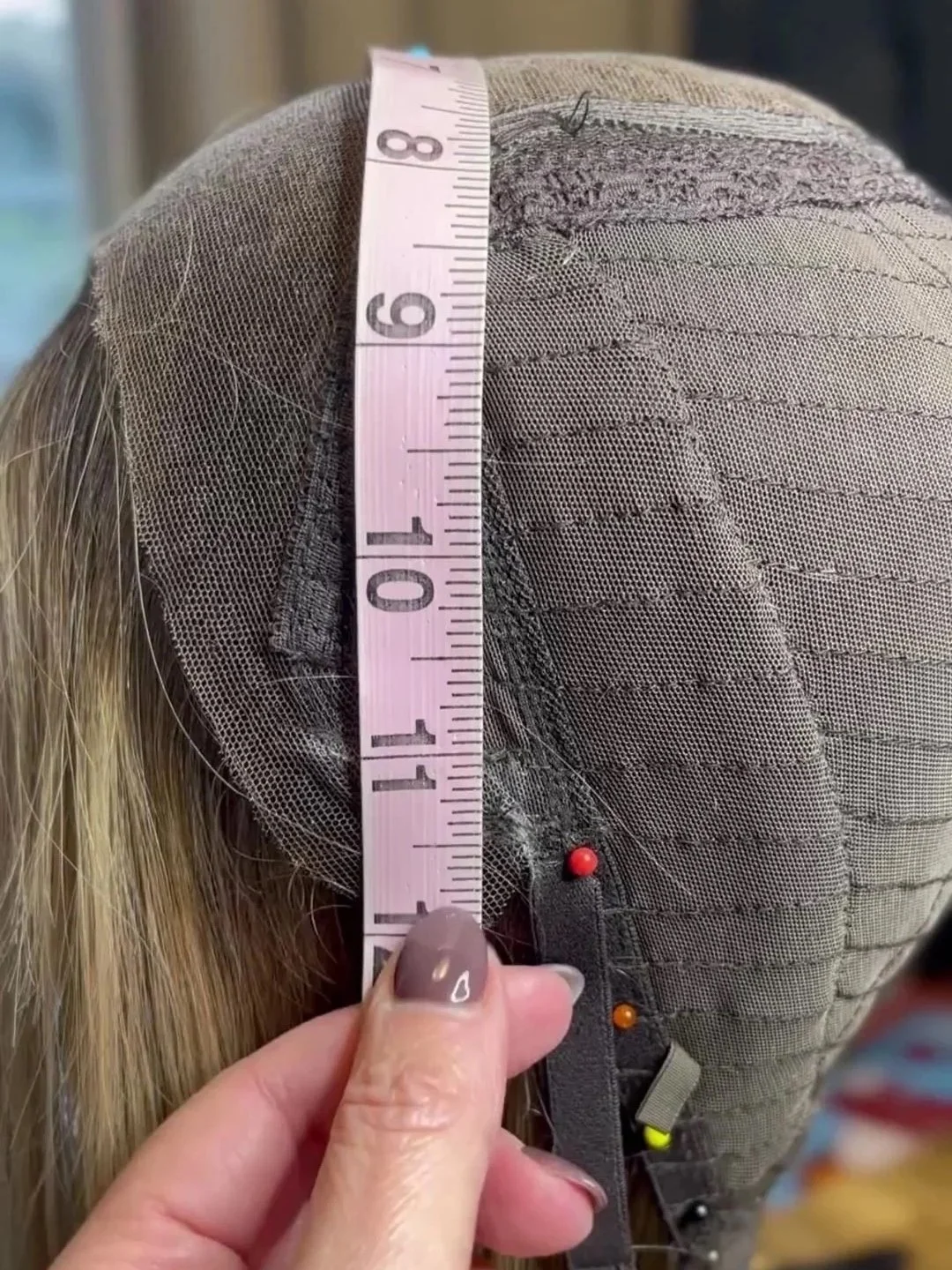 Close-up of a wig cap being measured with a pink measuring tape, showing a head covering with fine netting and some pins.