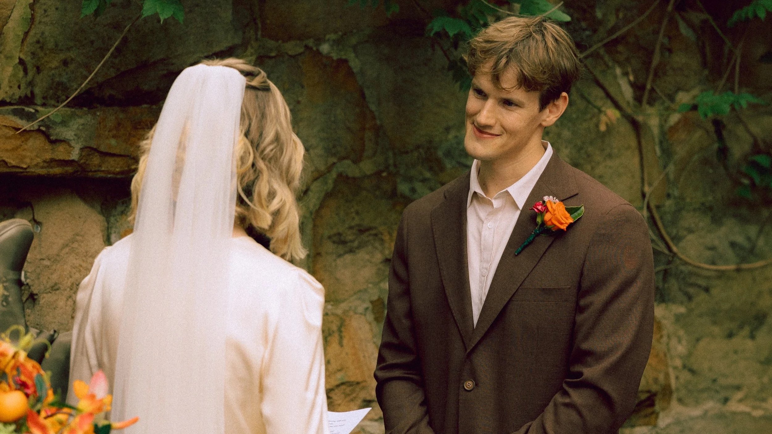 A young man and woman standing indoors in front of a stone wall, during a wedding ceremony. The man is wearing a brown suit with an orange and pink boutonniere. The woman is wearing a white wedding dress and veil, and holds a piece of paper.