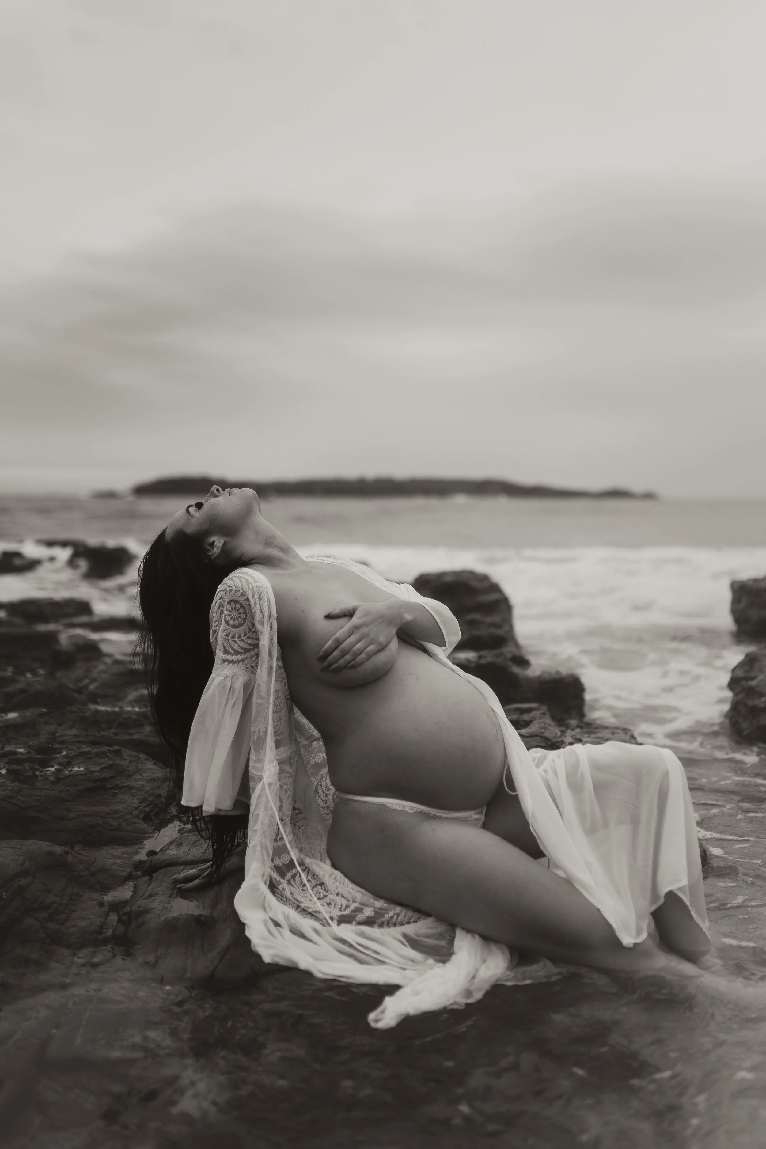 Black and white photo of a pregnant woman wearing a lace robe, sitting on rocks by the shoreline with her head tilted back and eyes closed.