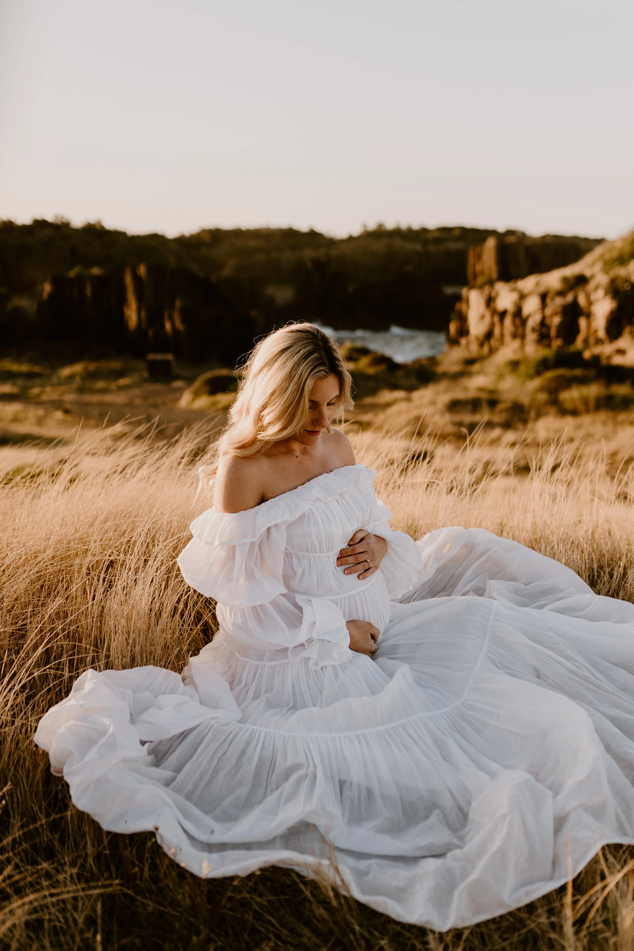 A pregnant woman in a white off-the-shoulder dress sitting on grassy field during golden hour, with rocky cliffs and ocean in the background.