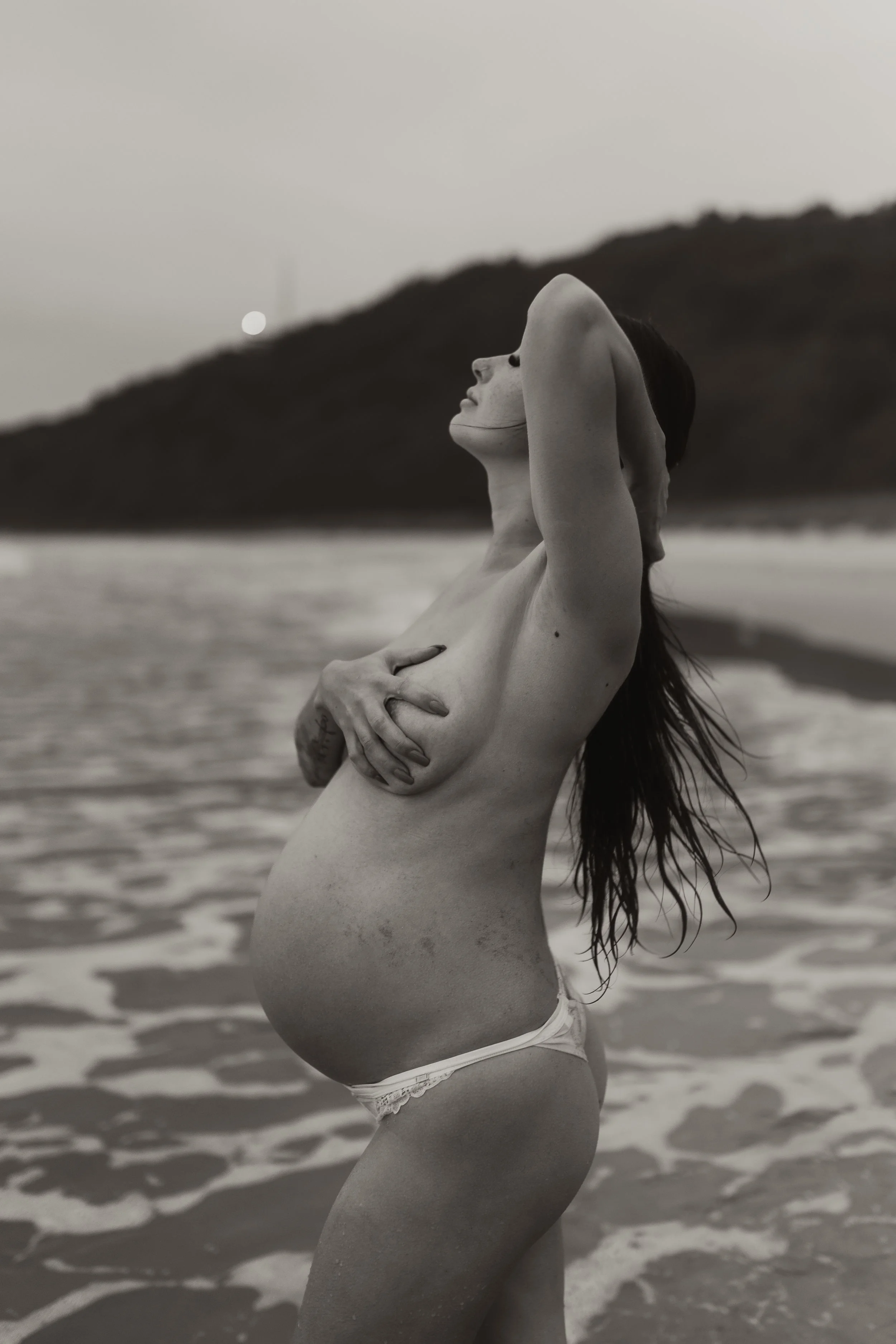 A pregnant woman standing in the ocean holding her chest with her eyes closed and head tilted back.