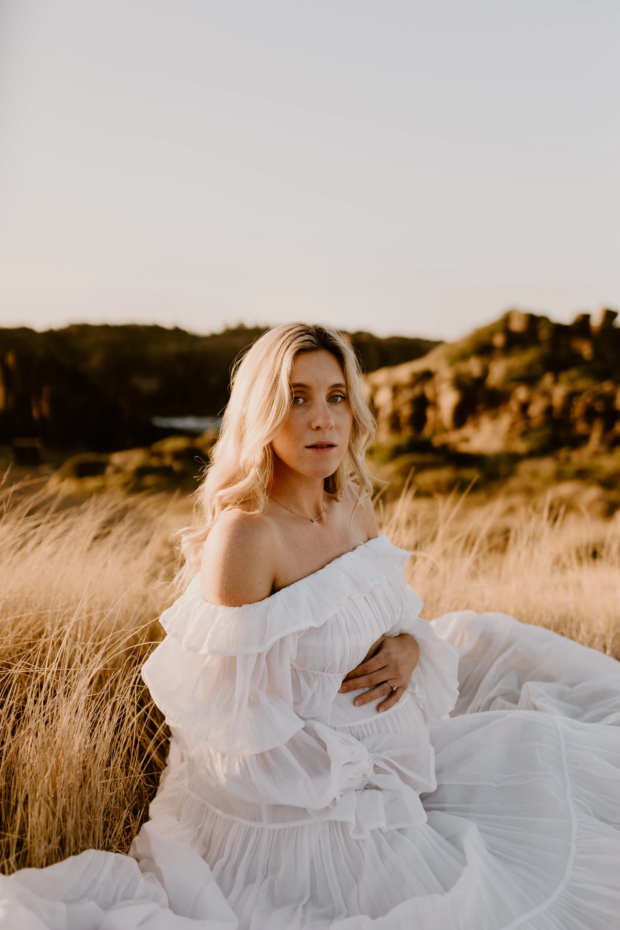 A woman with blonde hair sitting in a field of tall, golden grass, wearing a white off-shoulder dress, during sunset in a natural landscape with cliffs in the background.