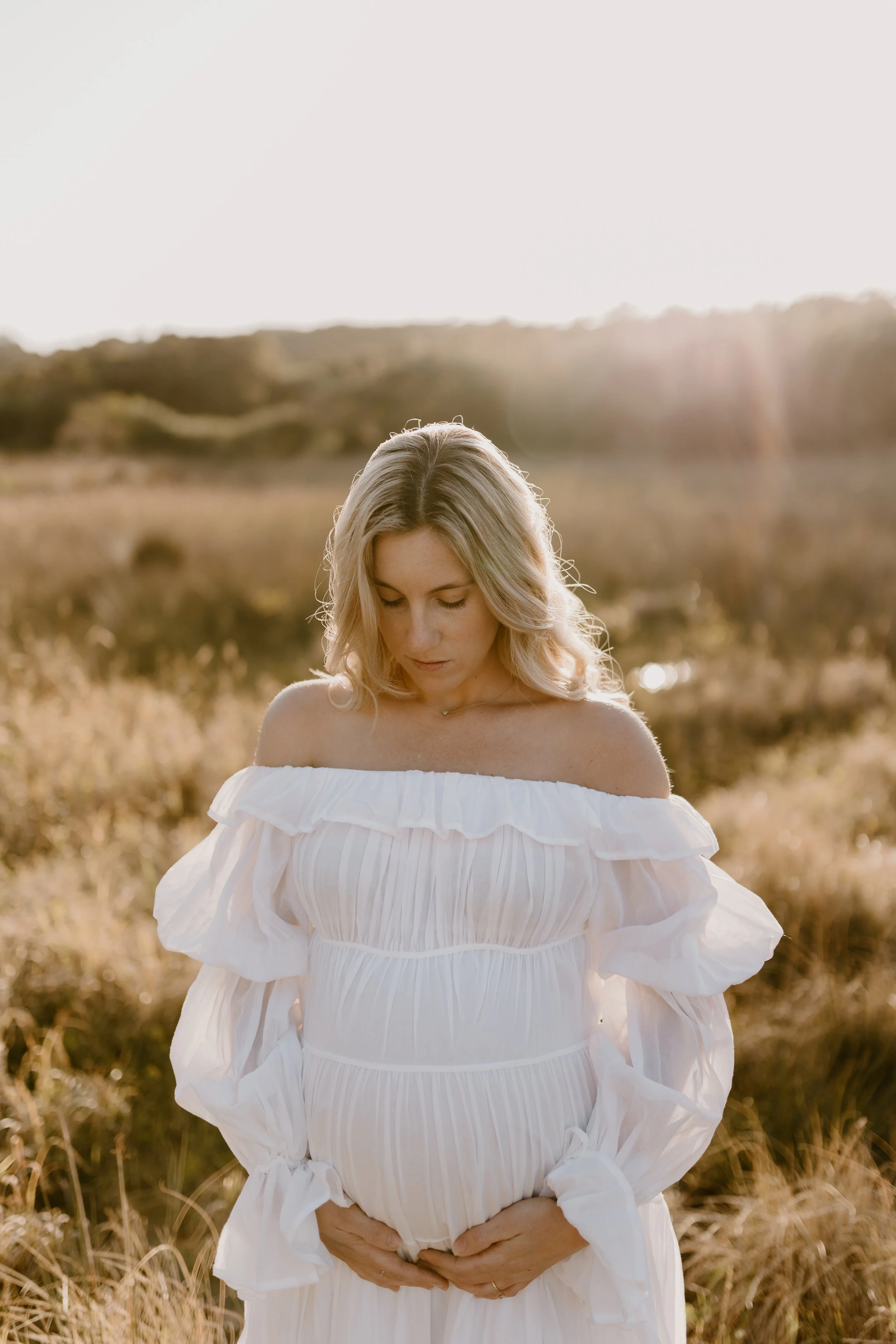 A pregnant woman in a white off-shoulder dress standing in a grassy field during sunset.