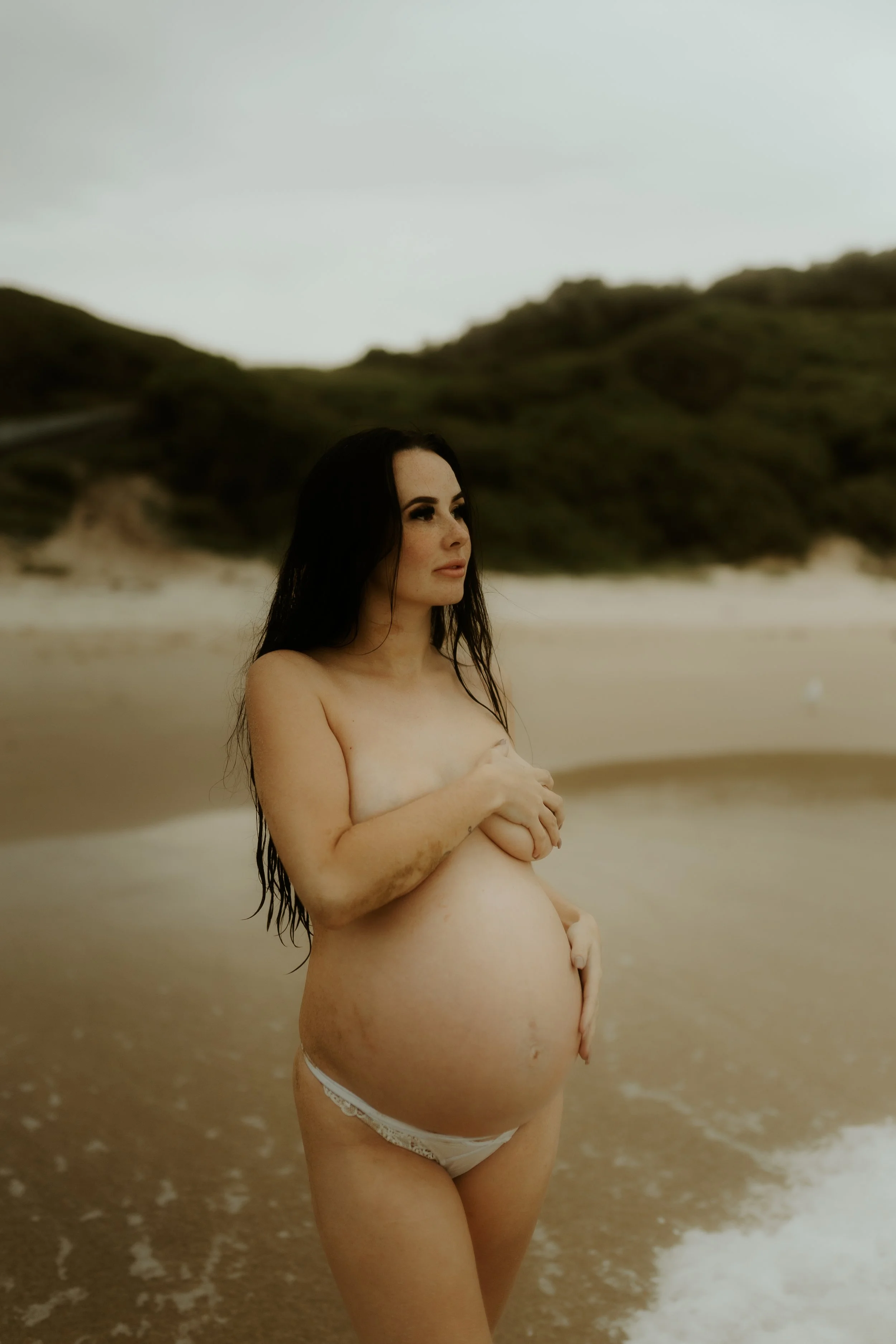 Pregnant woman standing on a beach, holding her exposed belly and covering her breasts with her hands, with a background of hills and ocean.
