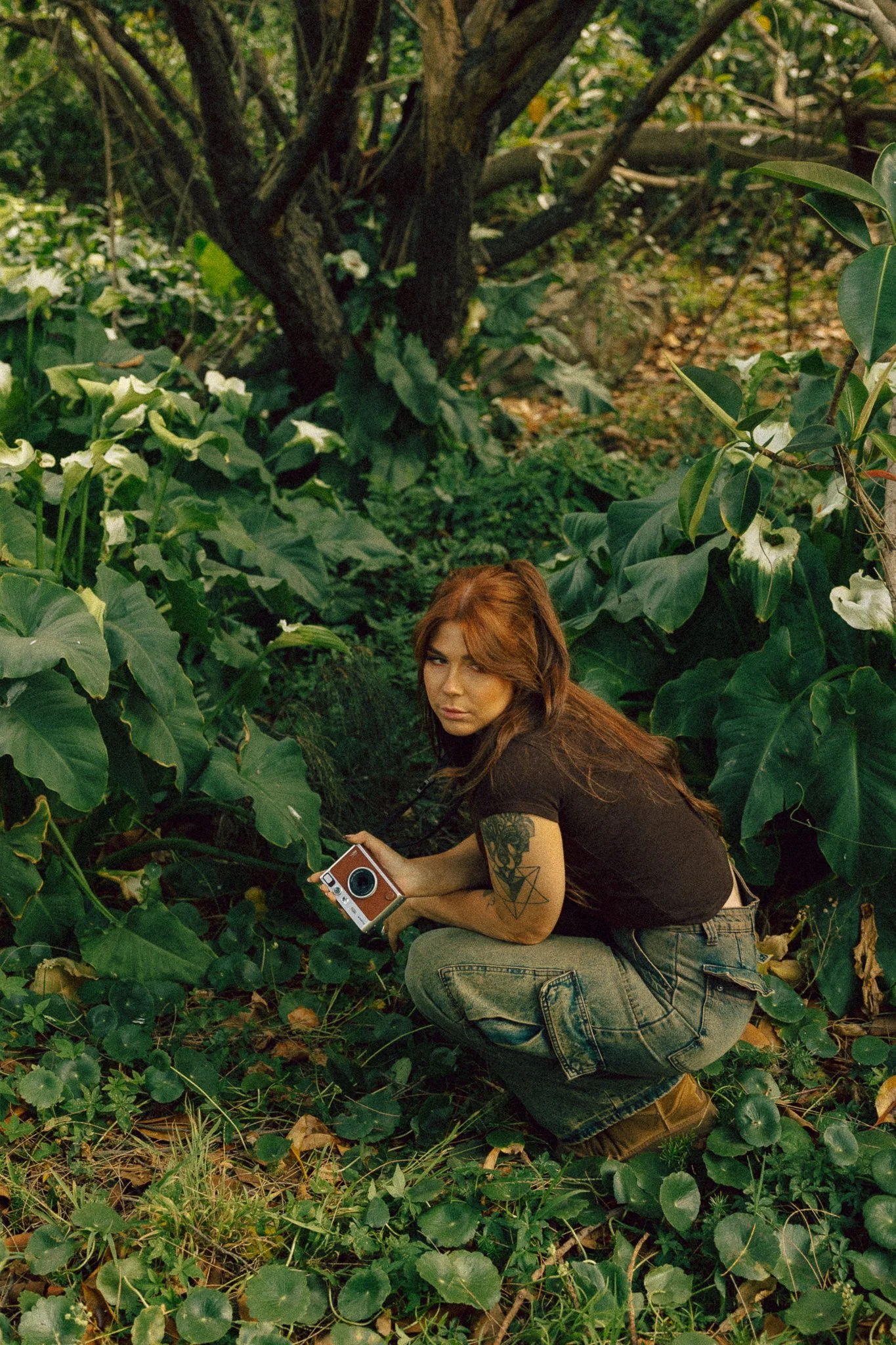 A woman with long brown hair, tattoos on her arm, wearing a black shirt and gray jeans, crouching in a dense, green forest surrounded by large leafy plants, holding a brown and white camera.