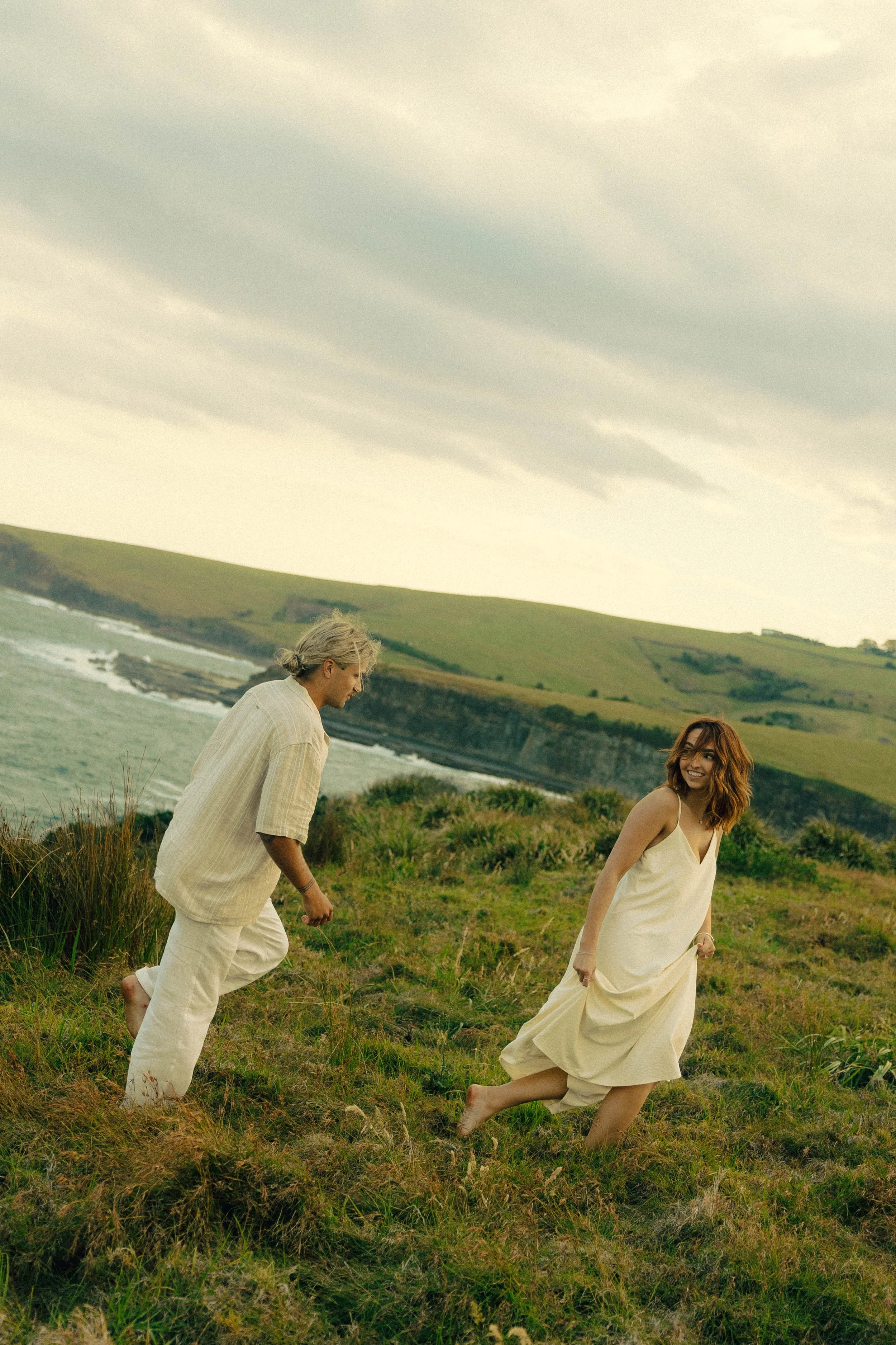 A young woman with reddish hair in a cream dress smiling, running barefoot on a grassy hill by the coast, with an older man with gray hair in light-colored clothes, also barefoot, following her.