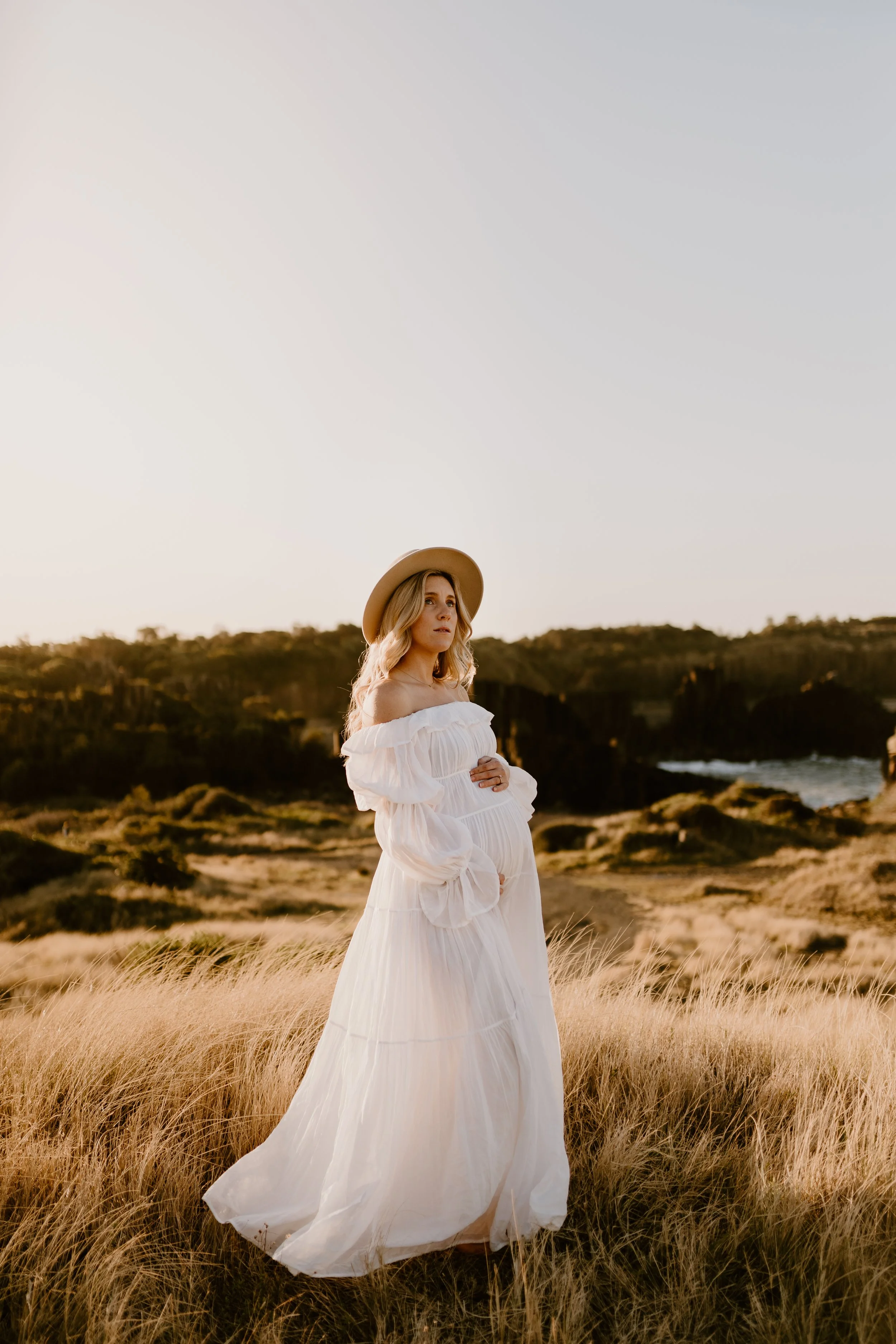 A pregnant woman in a flowing white dress and a wide-brimmed hat, standing in a grassy field during sunset with hills and the ocean in the background.