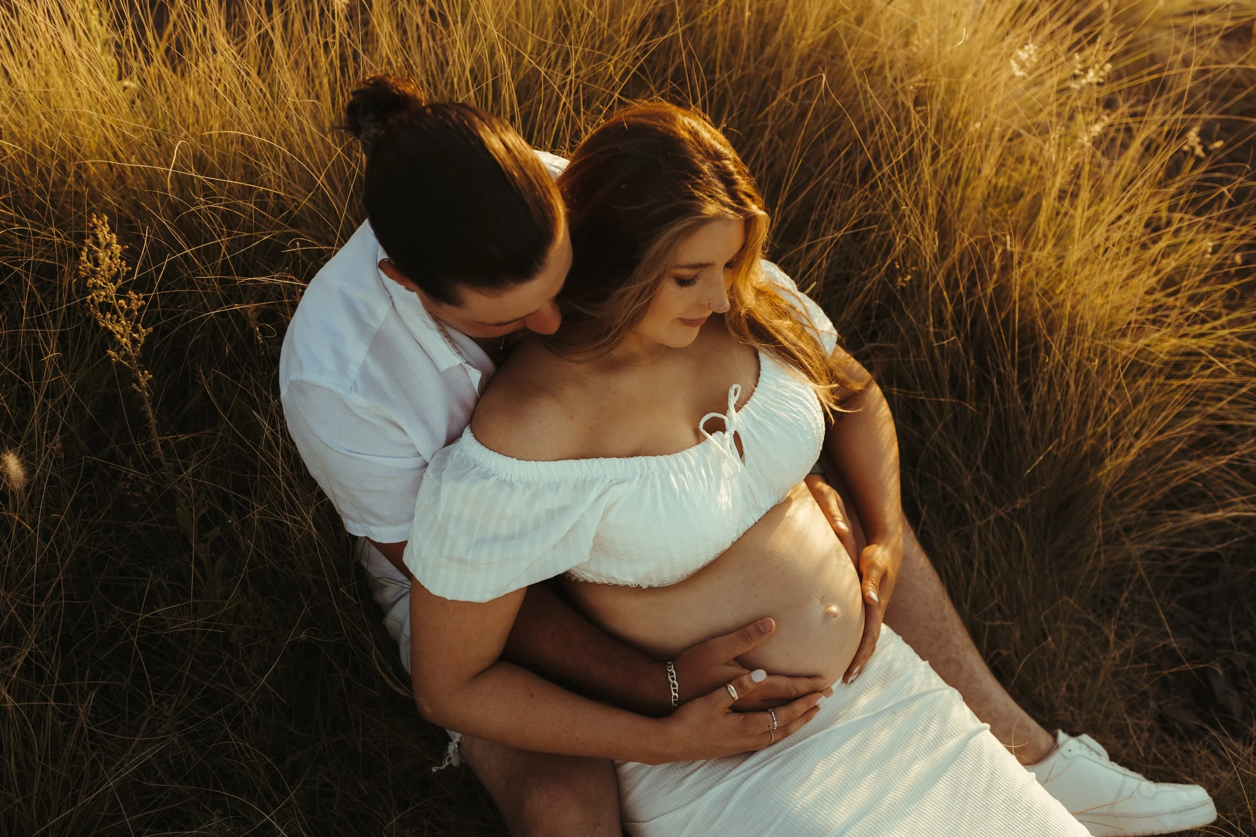 A couple sitting together in a grassy field, with the woman showing her pregnant belly and the man embracing her from behind during sunset.