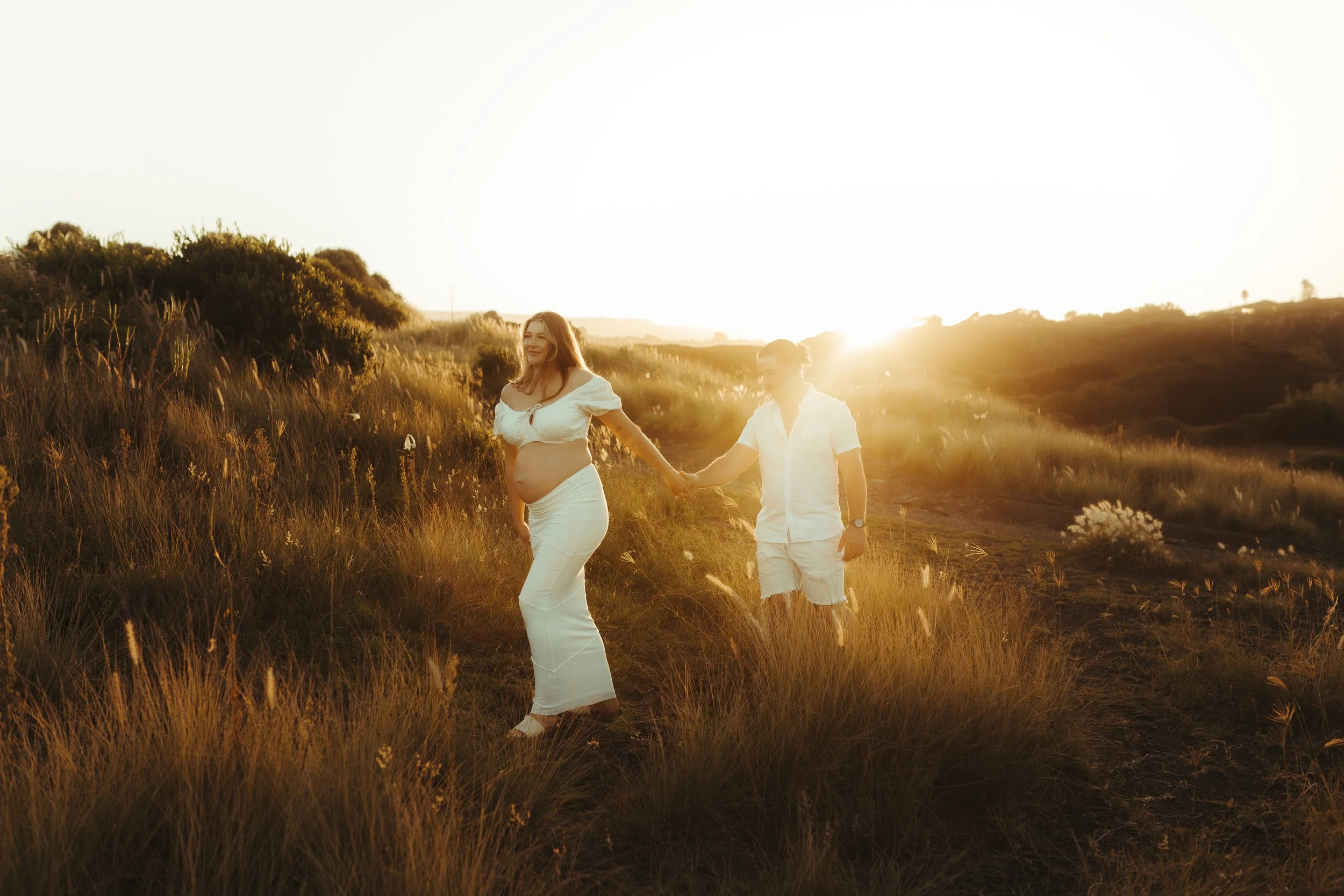 A pregnant woman and a man holding hands walking through a grassy field during sunset.