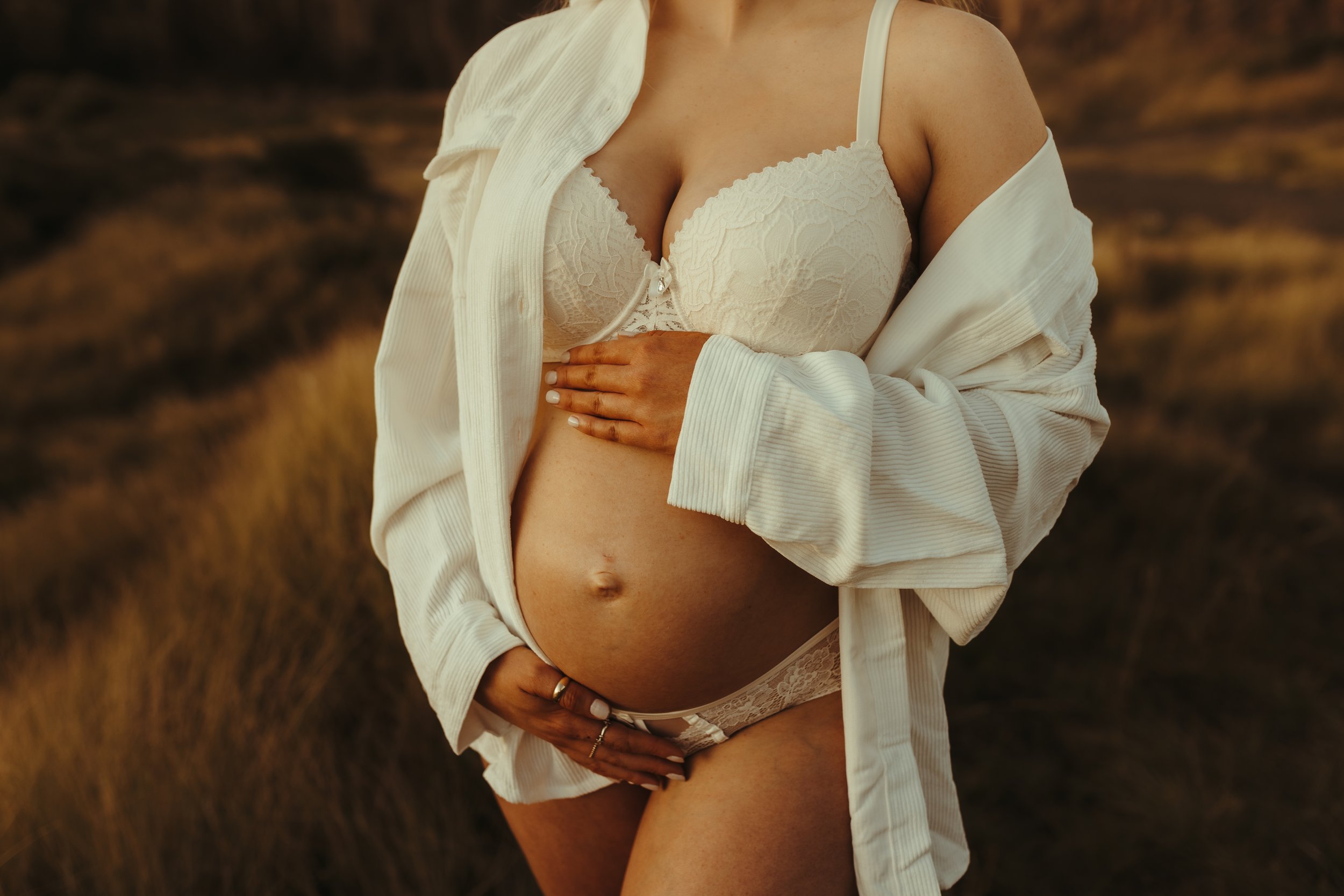 A pregnant woman wearing lace lingerie and an open white shirt, standing outdoors in a field during sunset.