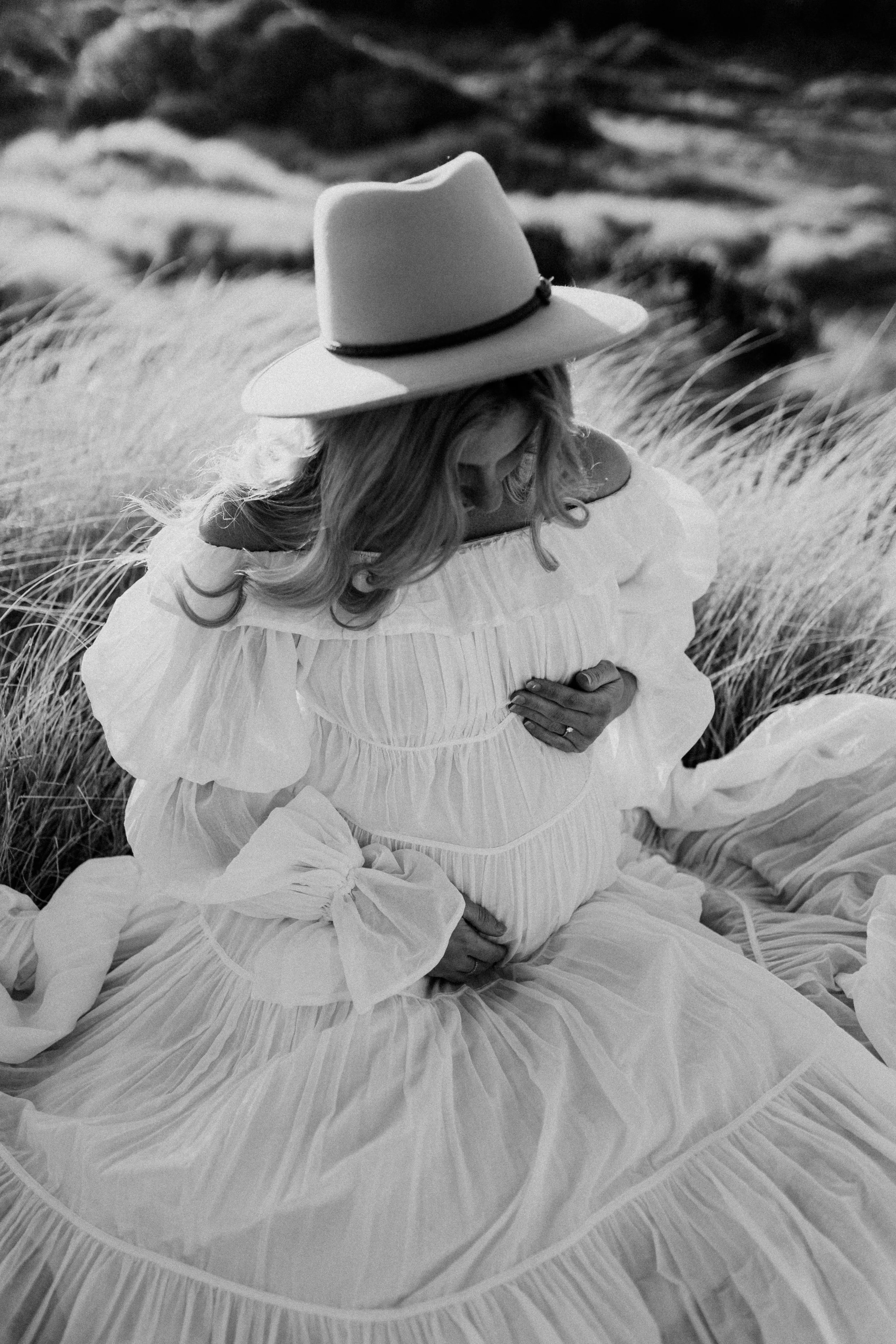 A woman in a flowing, off-the-shoulder dress with large bows sits on the grass in a field, wearing a wide-brimmed hat and looking down at her pregnant belly.