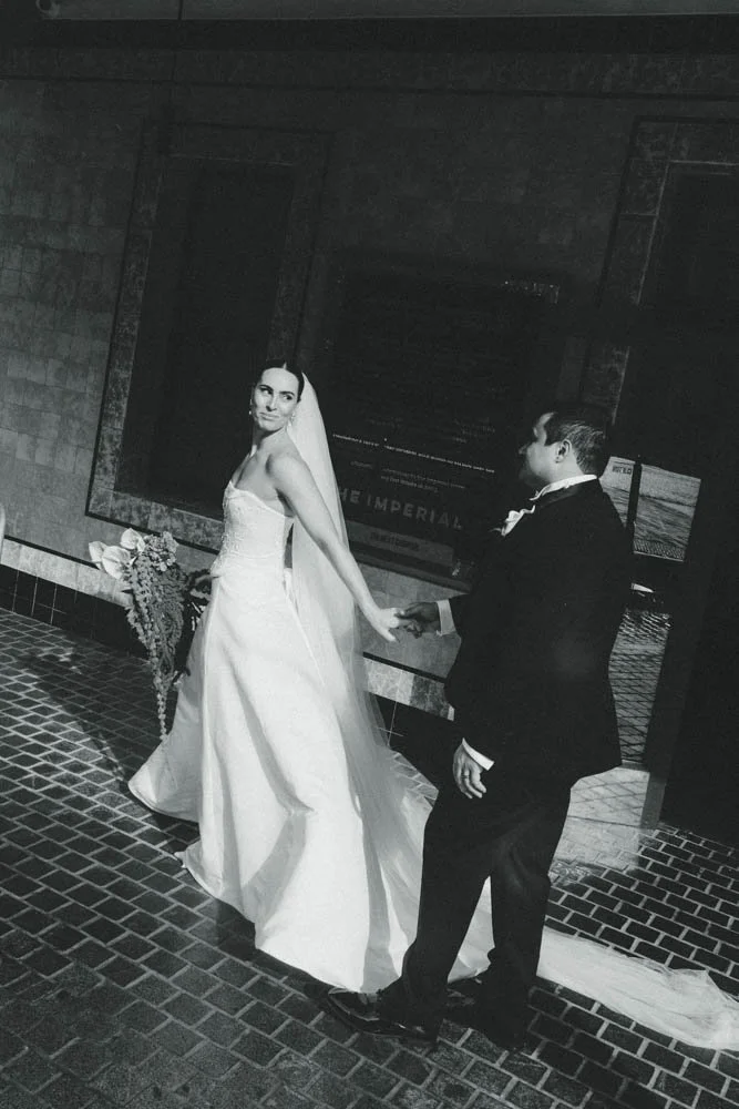 A black and white photo of a bride and groom holding hands, standing in front of a brick wall. The bride is in a wedding dress and veil, and the groom is in a tuxedo. A bouquet is placed on a bench beside them.