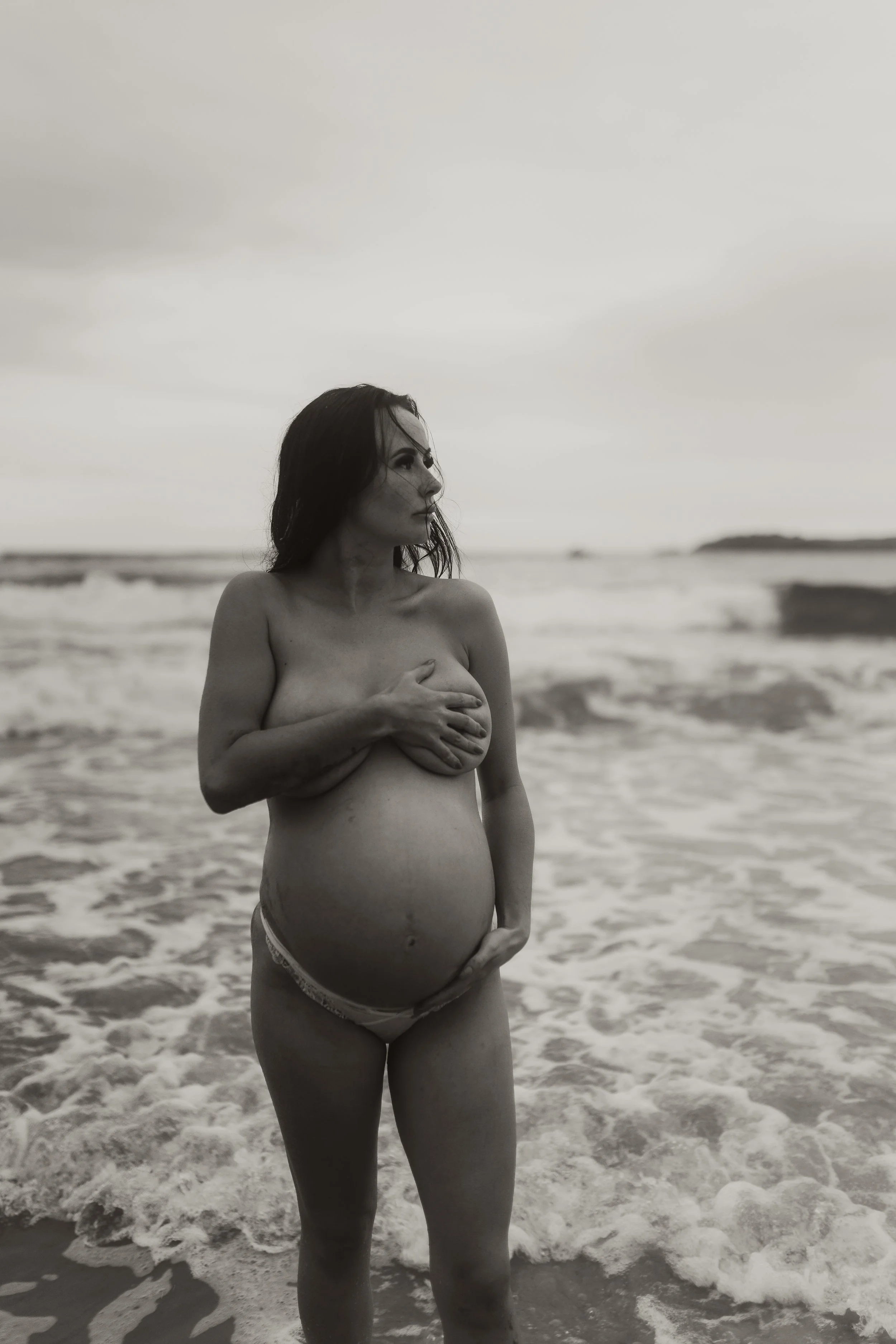 A pregnant woman stands in the ocean, holding her breast with one hand while the other hand rests on her belly. The photo is in black and white, with a cloudy sky in the background.